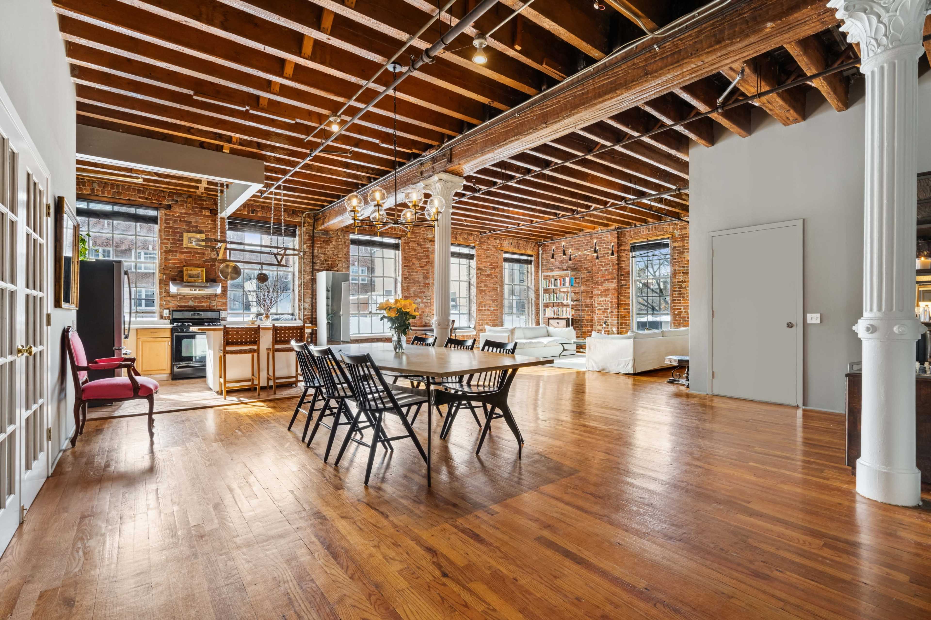 The image shows a spacious loft with exposed brick walls, wooden beams, and an open kitchen-dining area featuring a large table and seating.