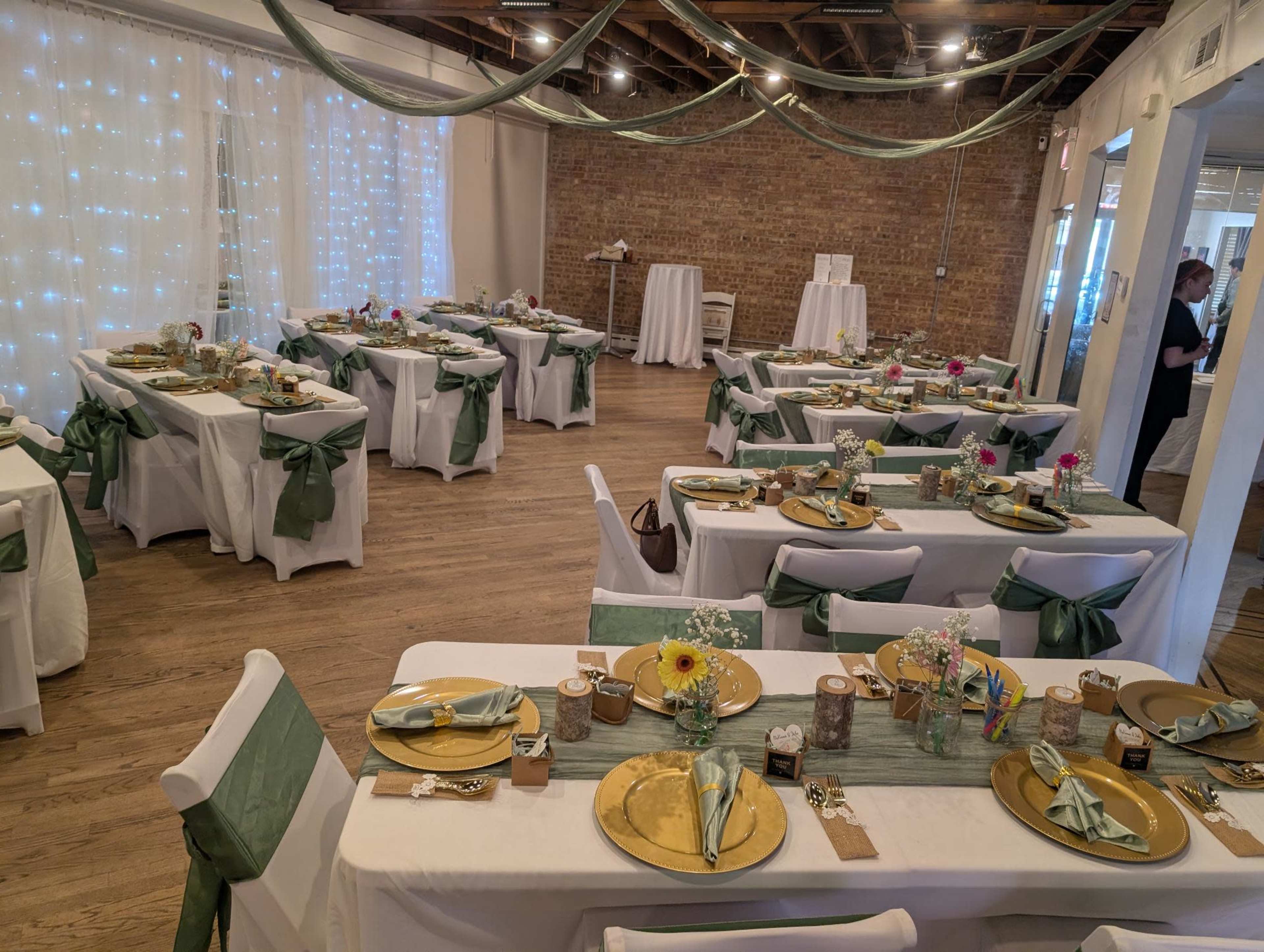 The image shows a decorated event venue with tables set for a banquet, featuring white tablecloths with green sashes, gold plates, and floral centerpieces.