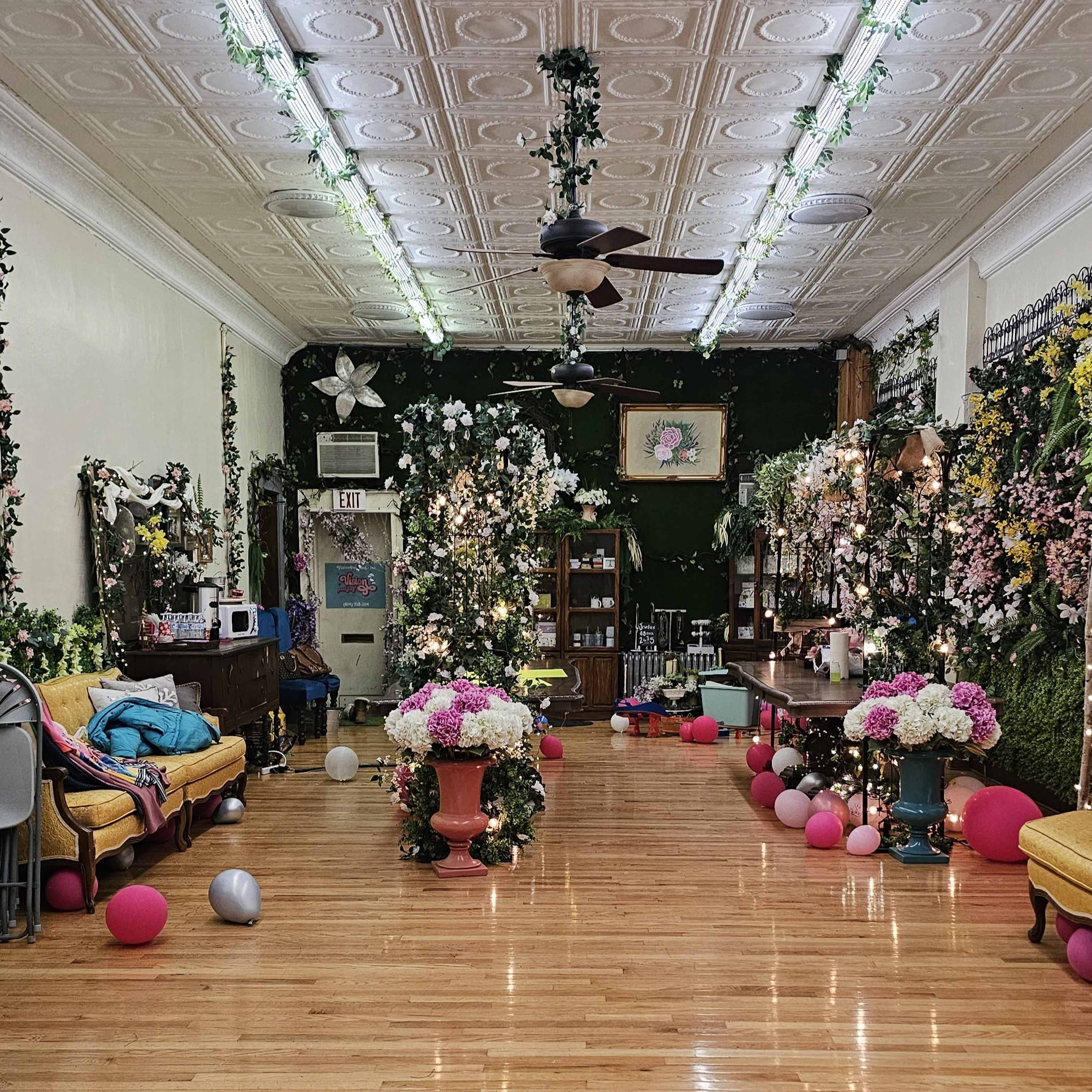 The image shows a decorated interior space with floral arrangements, balloons, and green foliage, arranged for a celebration or event.
