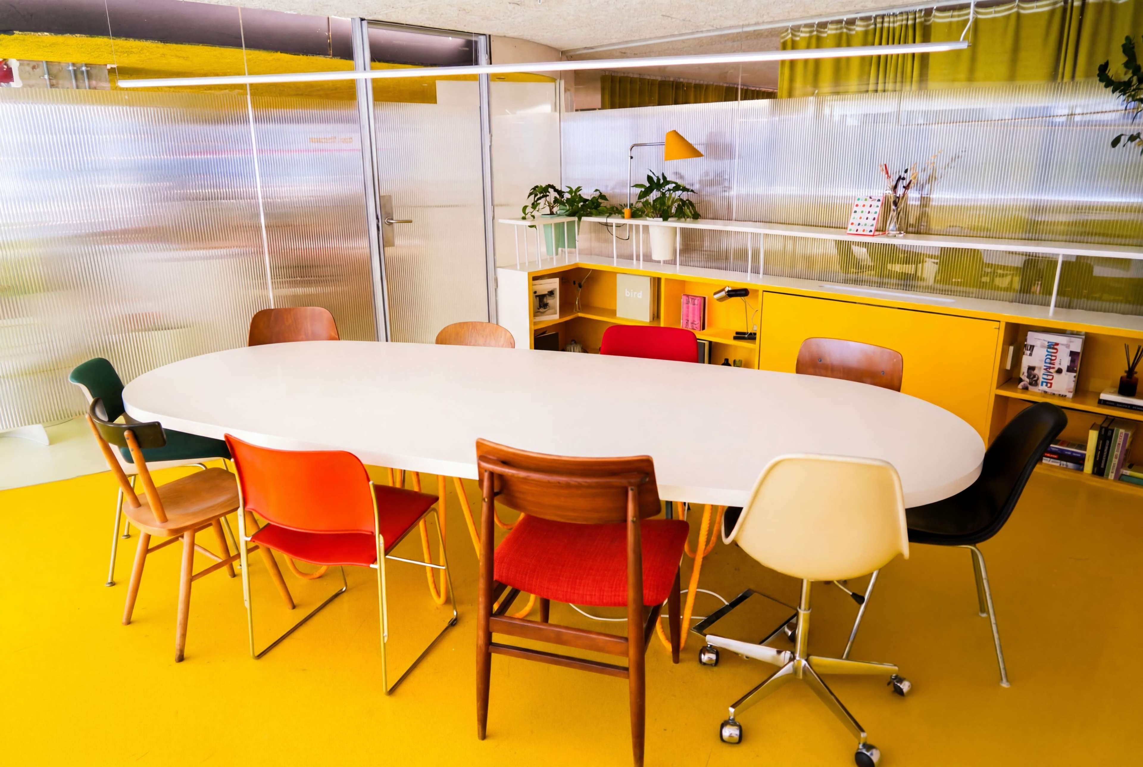 A modern meeting room features an oval table surrounded by eight chairs in various colors, with yellow flooring and glass partitions.