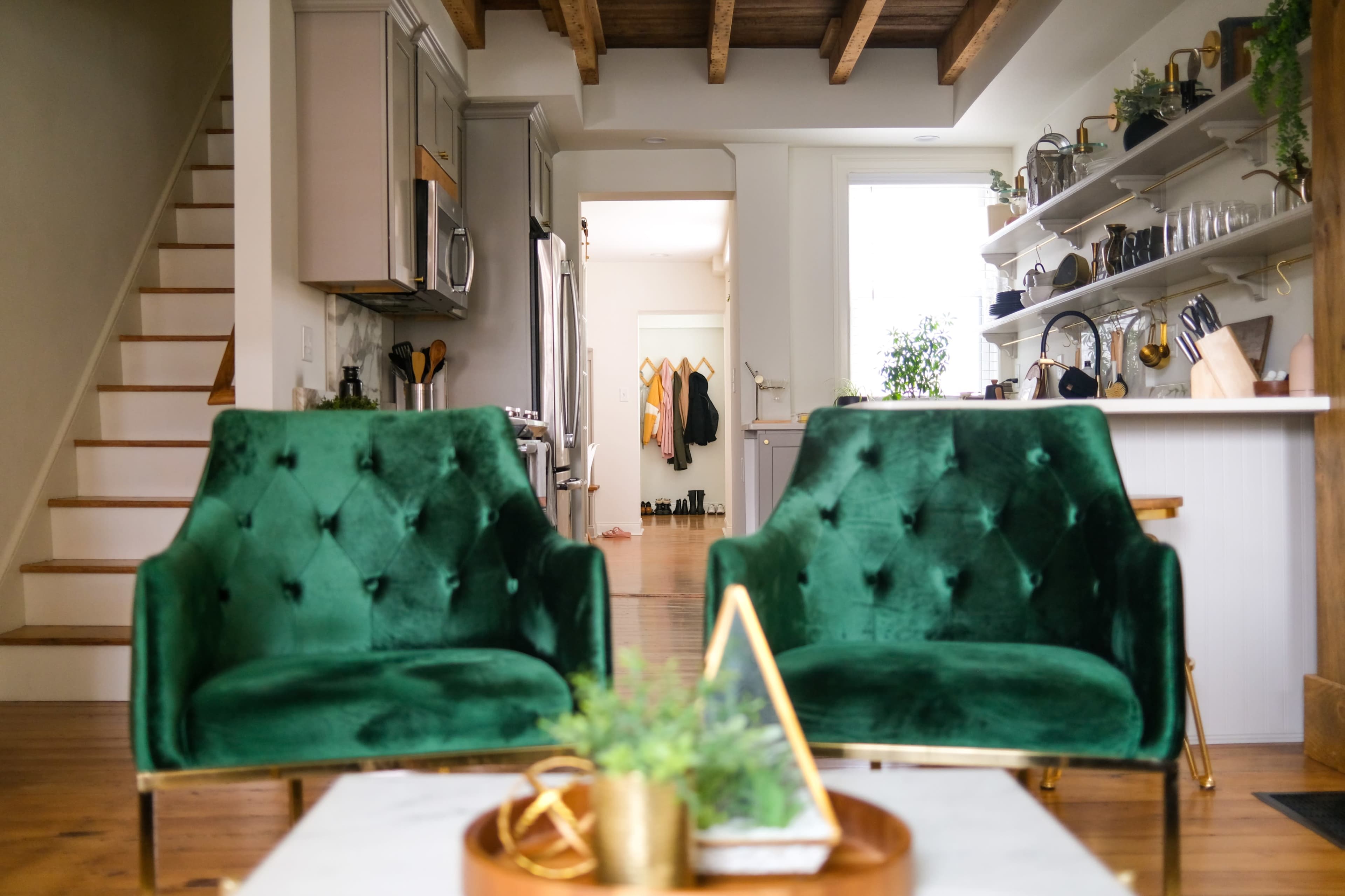 Two green velvet chairs facing a dining area with an open kitchen, shelves displaying kitchenware, and a staircase in the background.