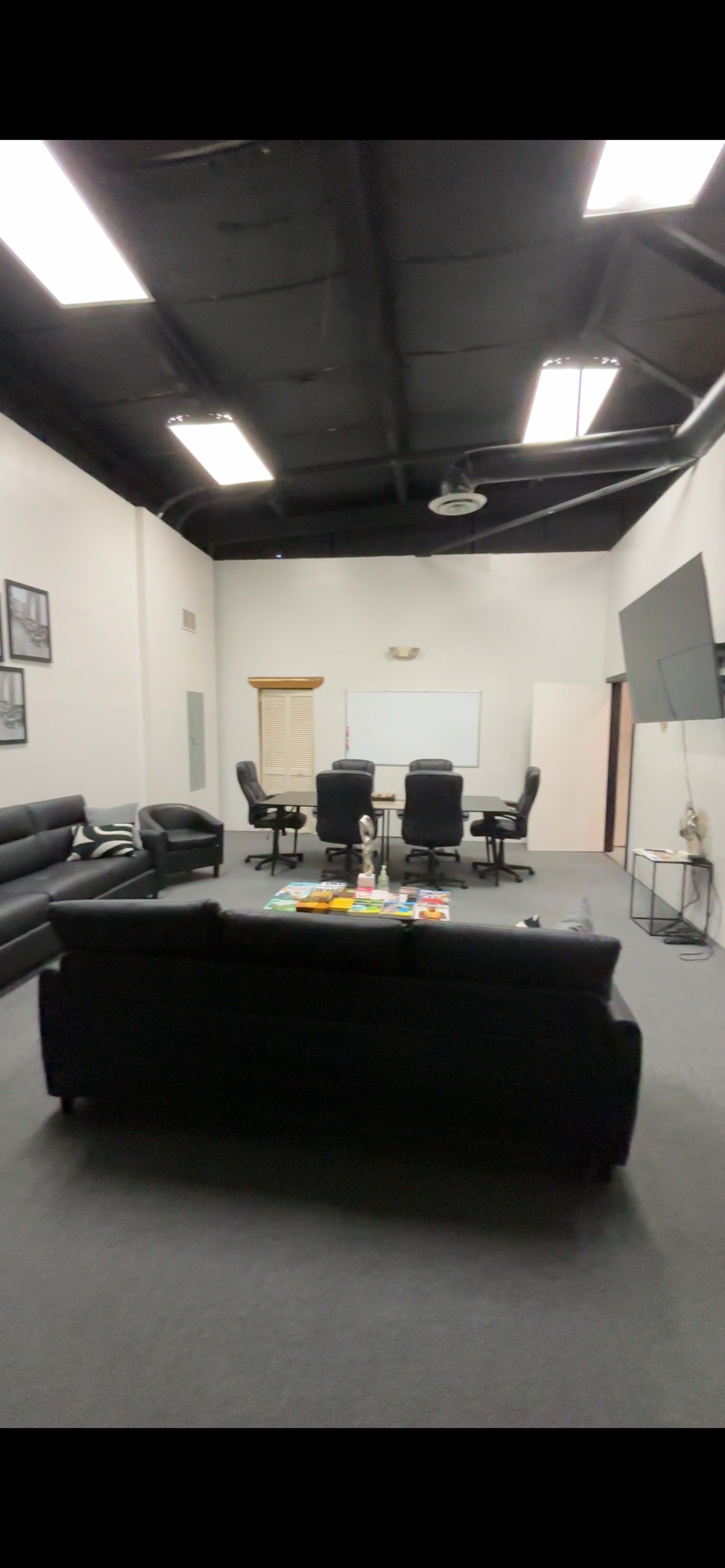 The image shows an office meeting room with black sofas, a table, several office chairs, and a large wall-mounted screen.