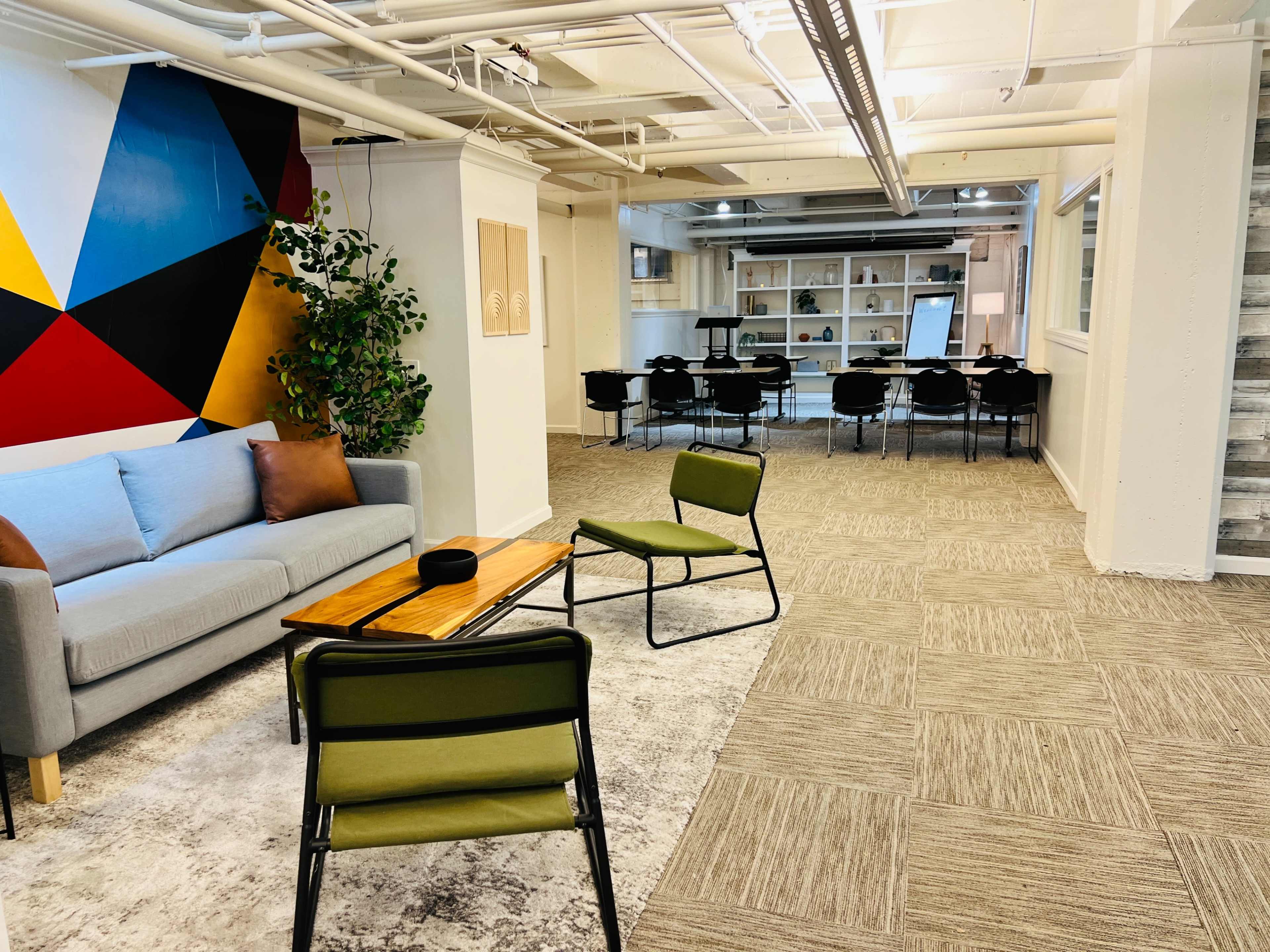 The image shows a modern workspace featuring a seating area with a gray sofa and two green chairs, leading to a dining area with a large table surrounded by black chairs.