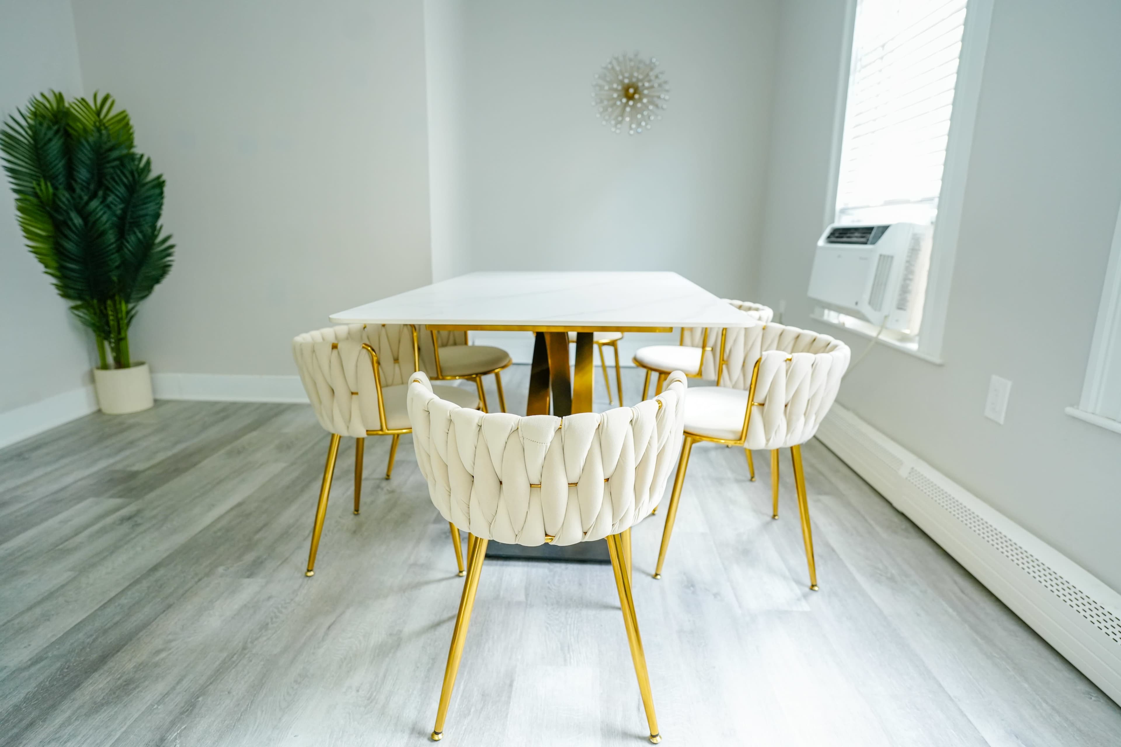 A modern dining room features a marble-top table surrounded by four upholstered chairs with gold legs, with a potted plant in the corner and an air conditioning unit by the window.