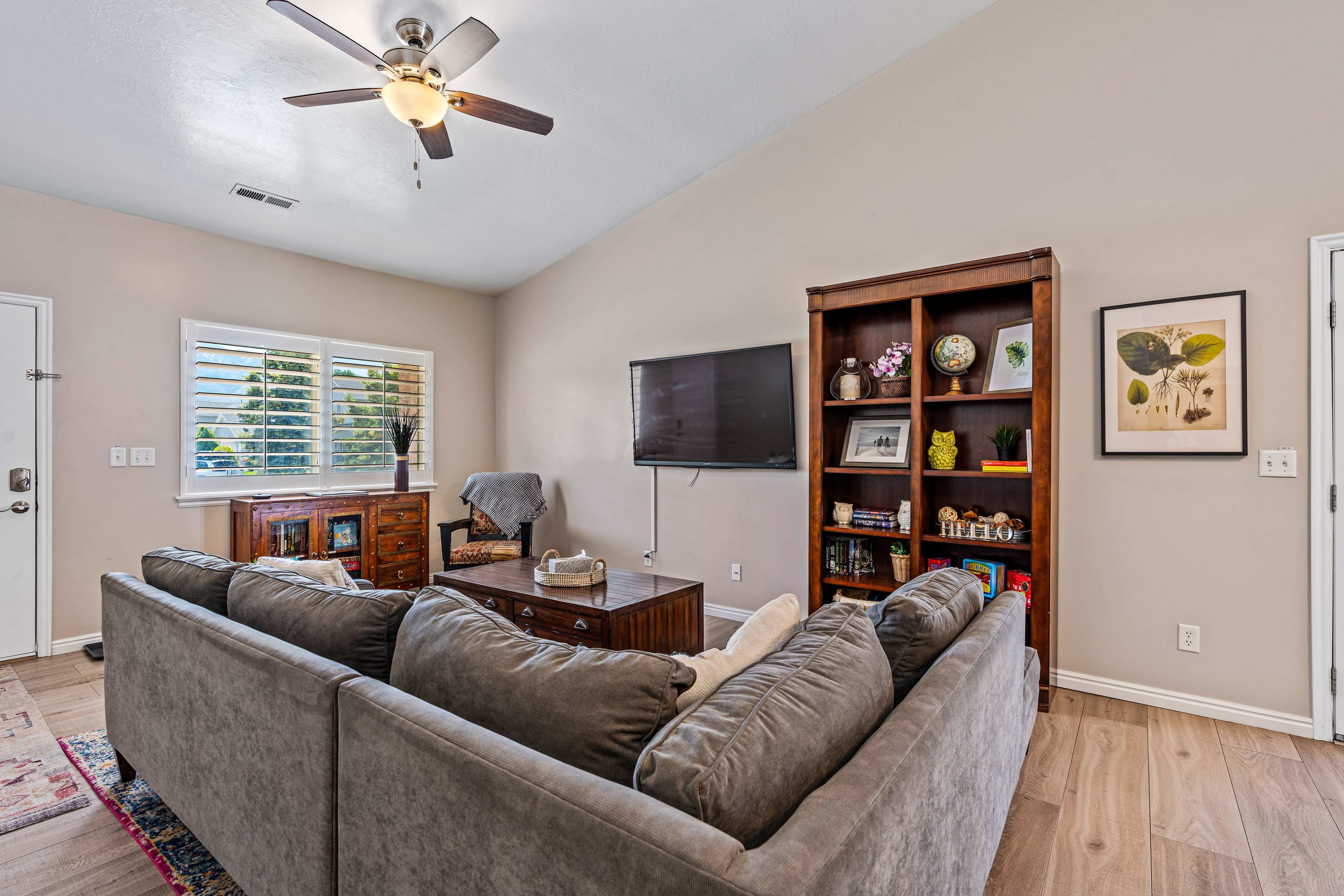 The living room features a gray sectional couch facing a mounted television on the wall, with a wooden bookshelf and a window on the opposite side.