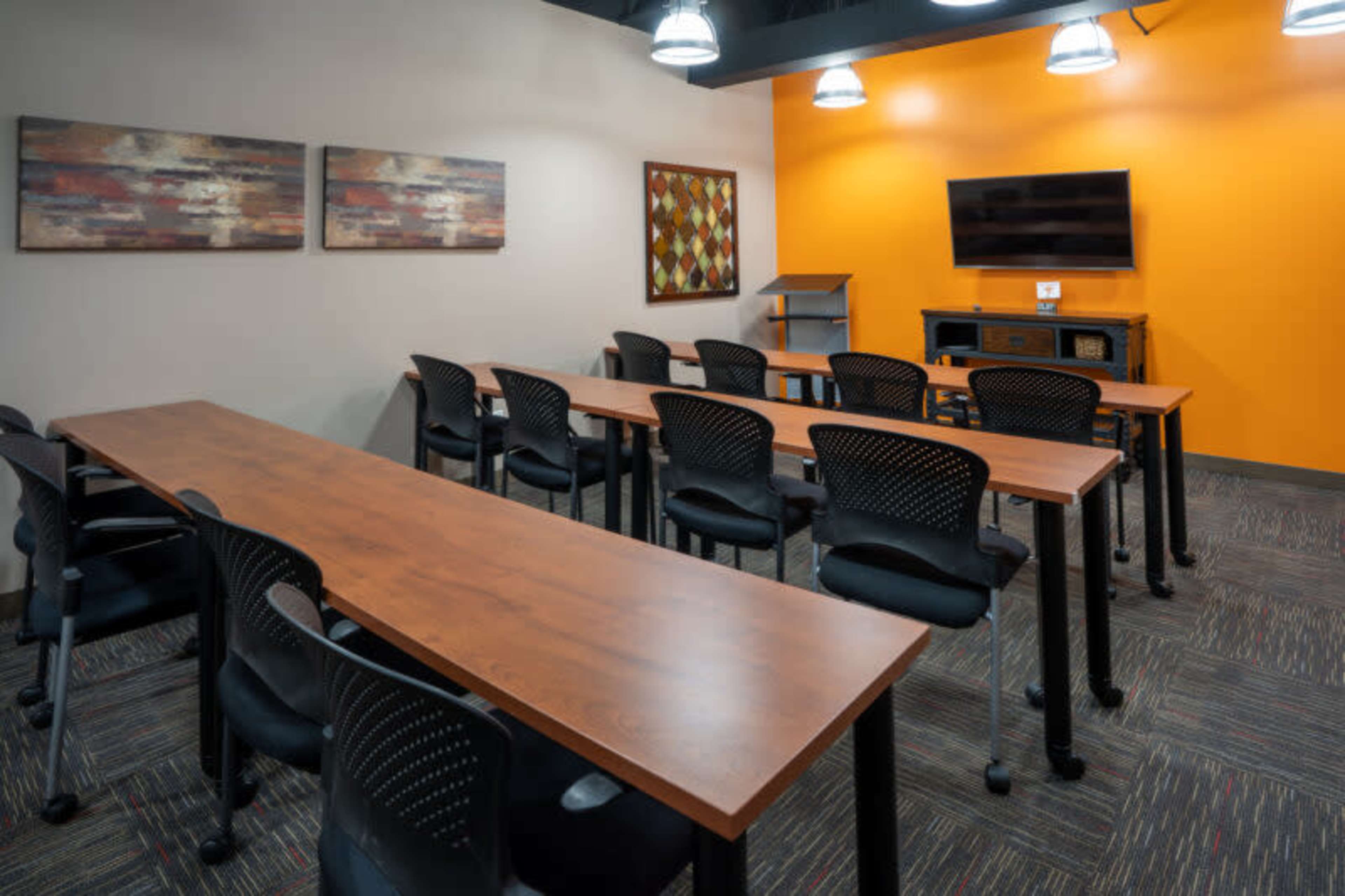 The image shows a modern meeting room with rows of wooden tables and chairs, an orange accent wall, and a television screen mounted on the wall.