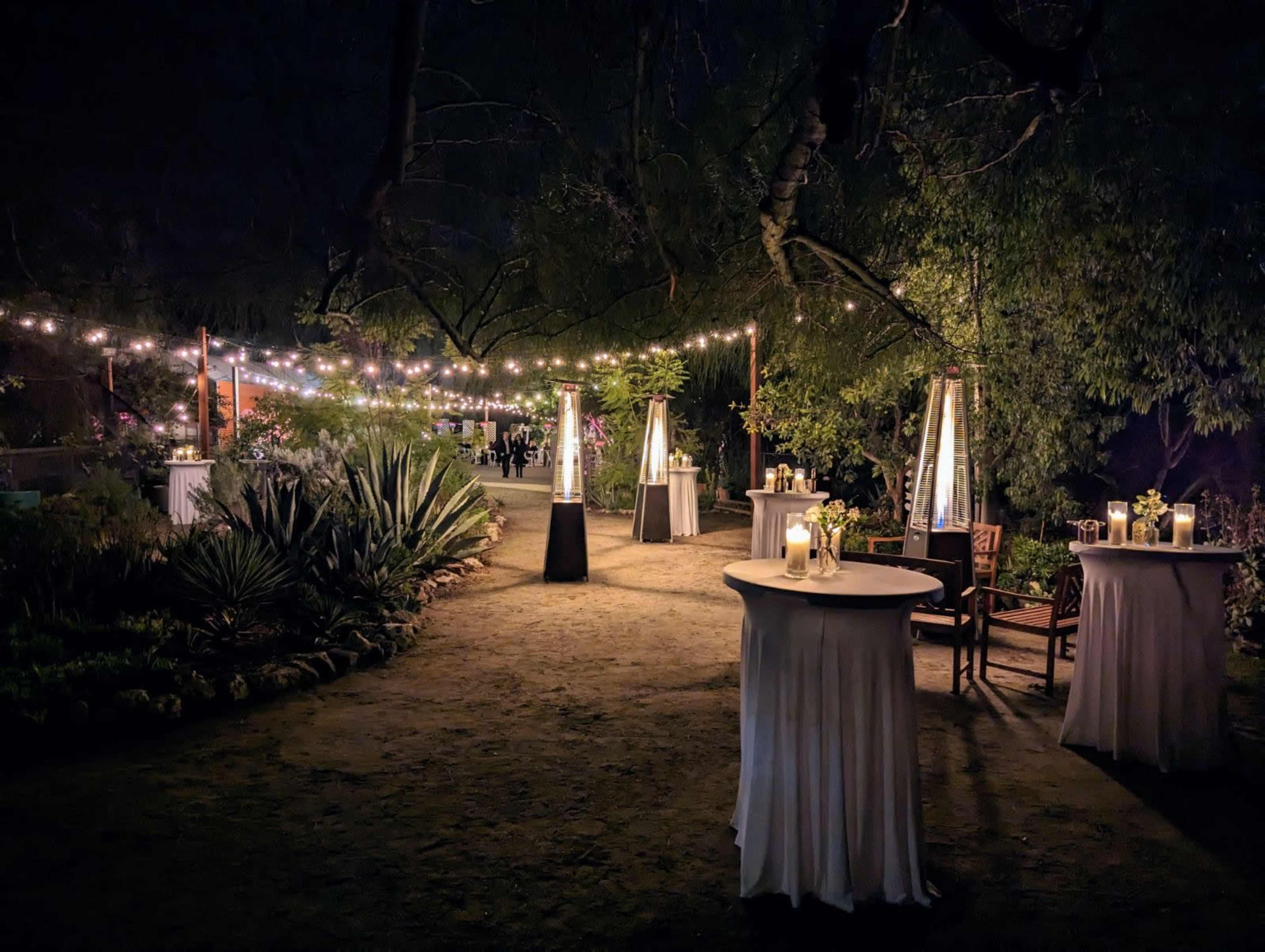 A beautifully lit outdoor event space with round tables and lanterns under strings of lights.