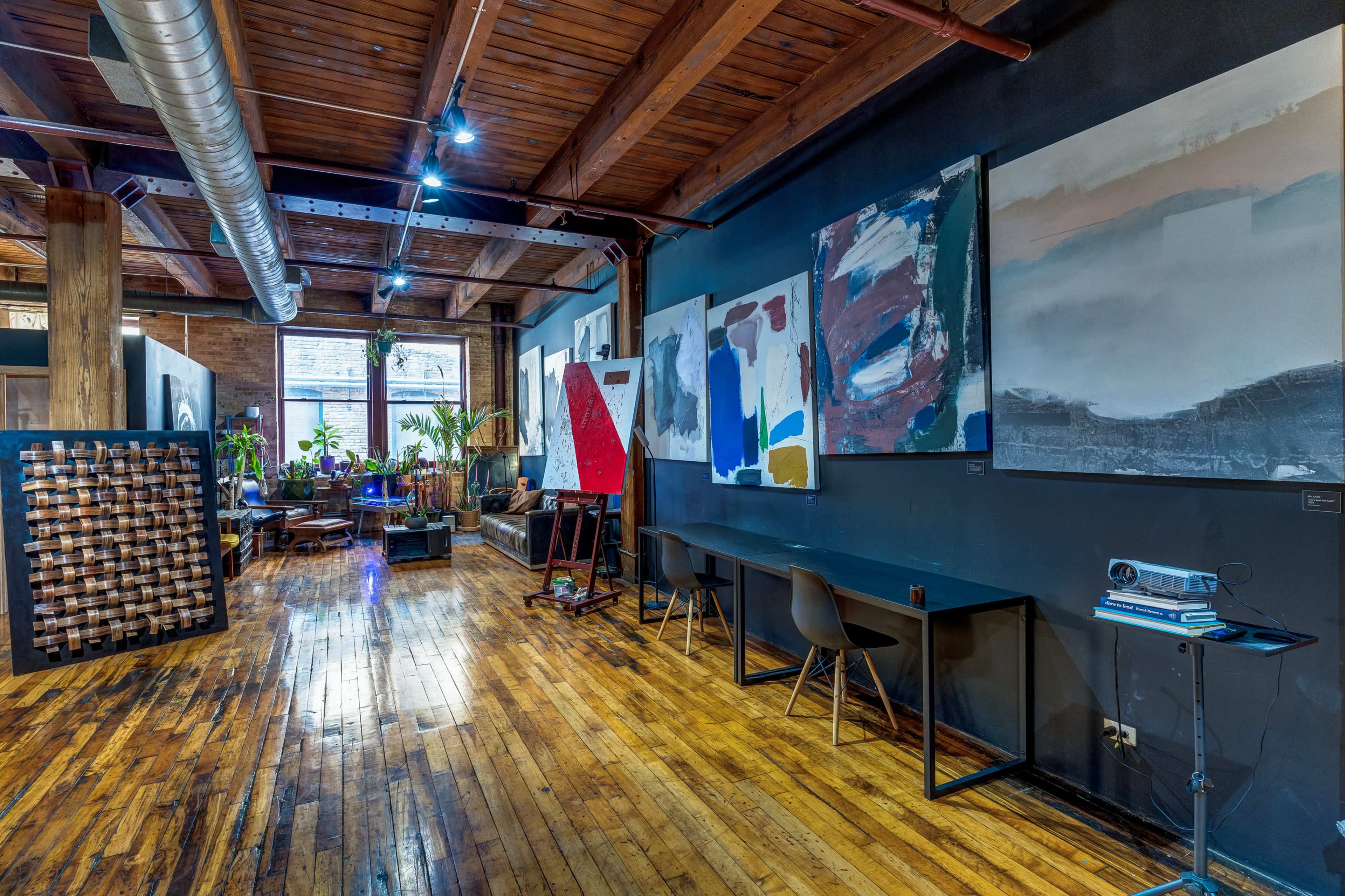 A spacious, industrial-style room with wooden floors, dark walls, and various large paintings hanging, along with a few seating areas and plants near a window.