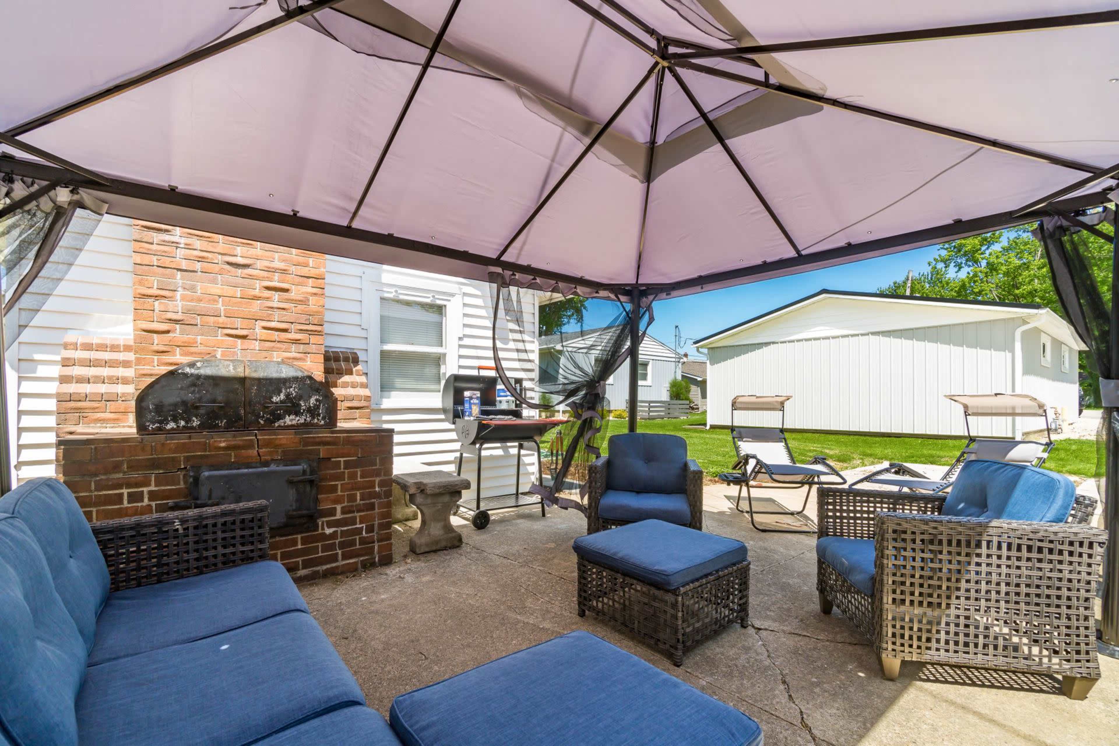 The image shows a shaded outdoor seating area with blue furniture, a brick barbecue grill, and a view of a lawn and nearby structures.