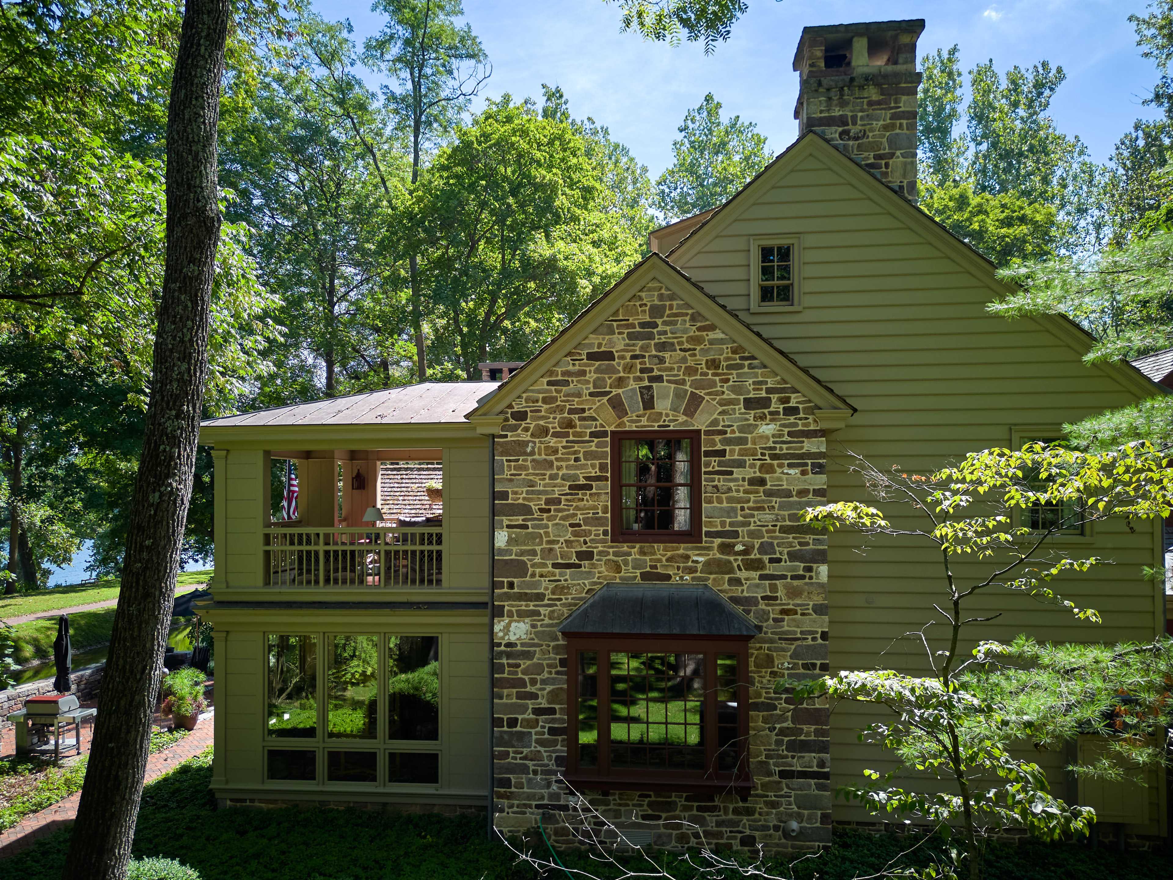 A two-story house featuring a stone facade on one side and wooden siding on the other, surrounded by lush trees and greenery.