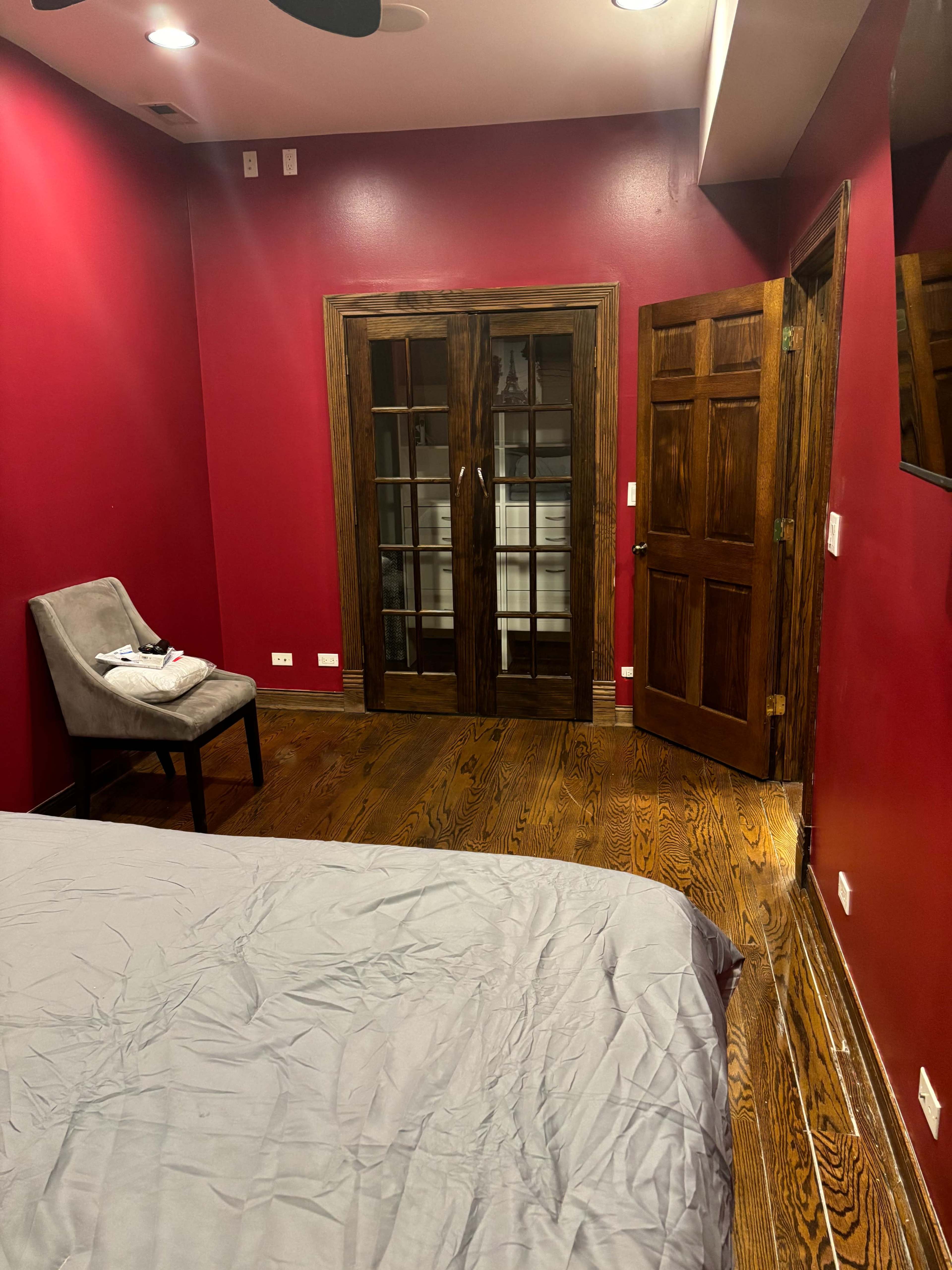 The image shows a bedroom with red walls, wooden flooring, a bed with gray bedding, a gray chair, and two doors leading to another room.