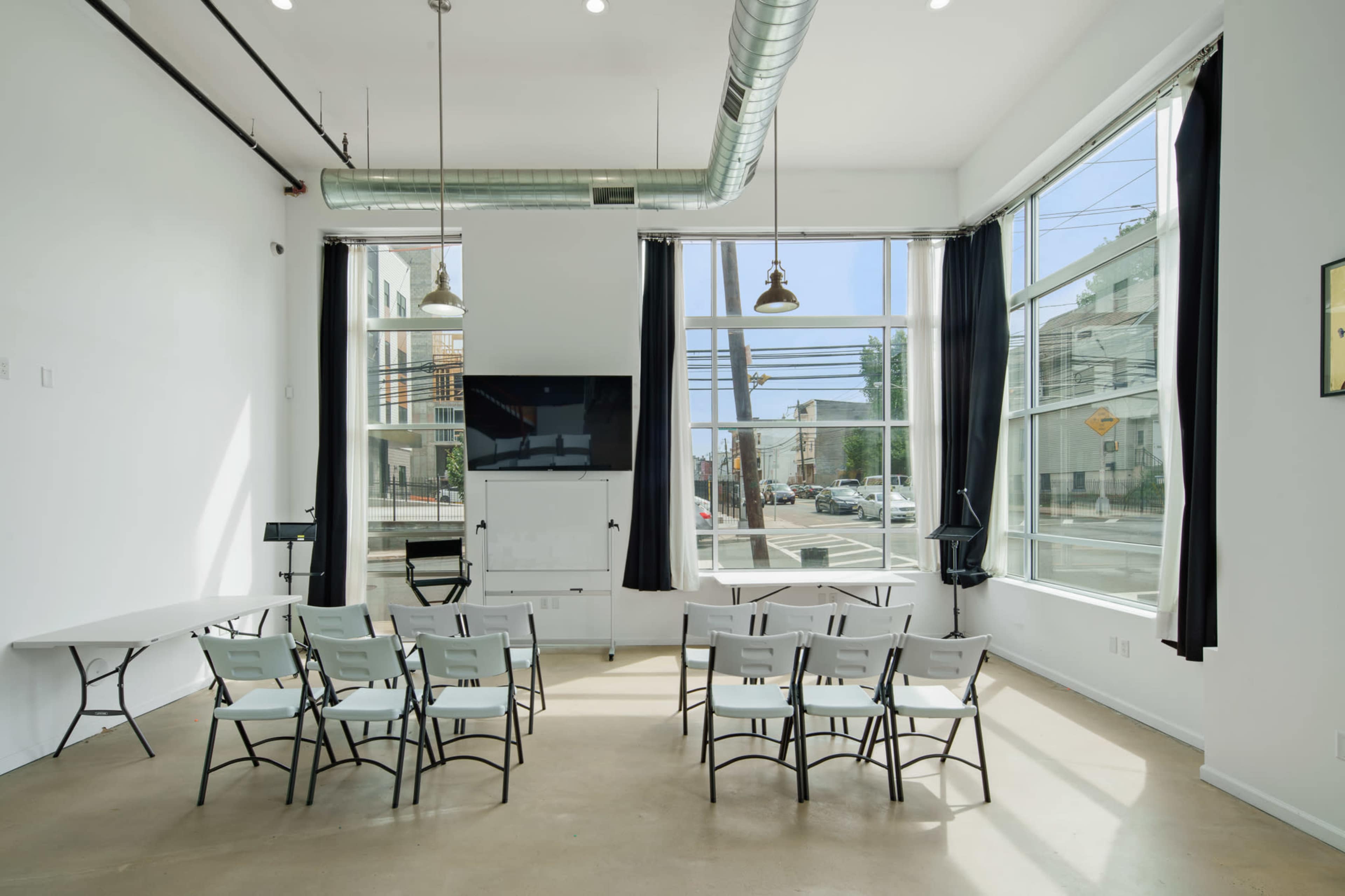 A bright, modern meeting room features several rows of metal chairs facing a presentation screen, with large windows allowing natural light to illuminate the space.