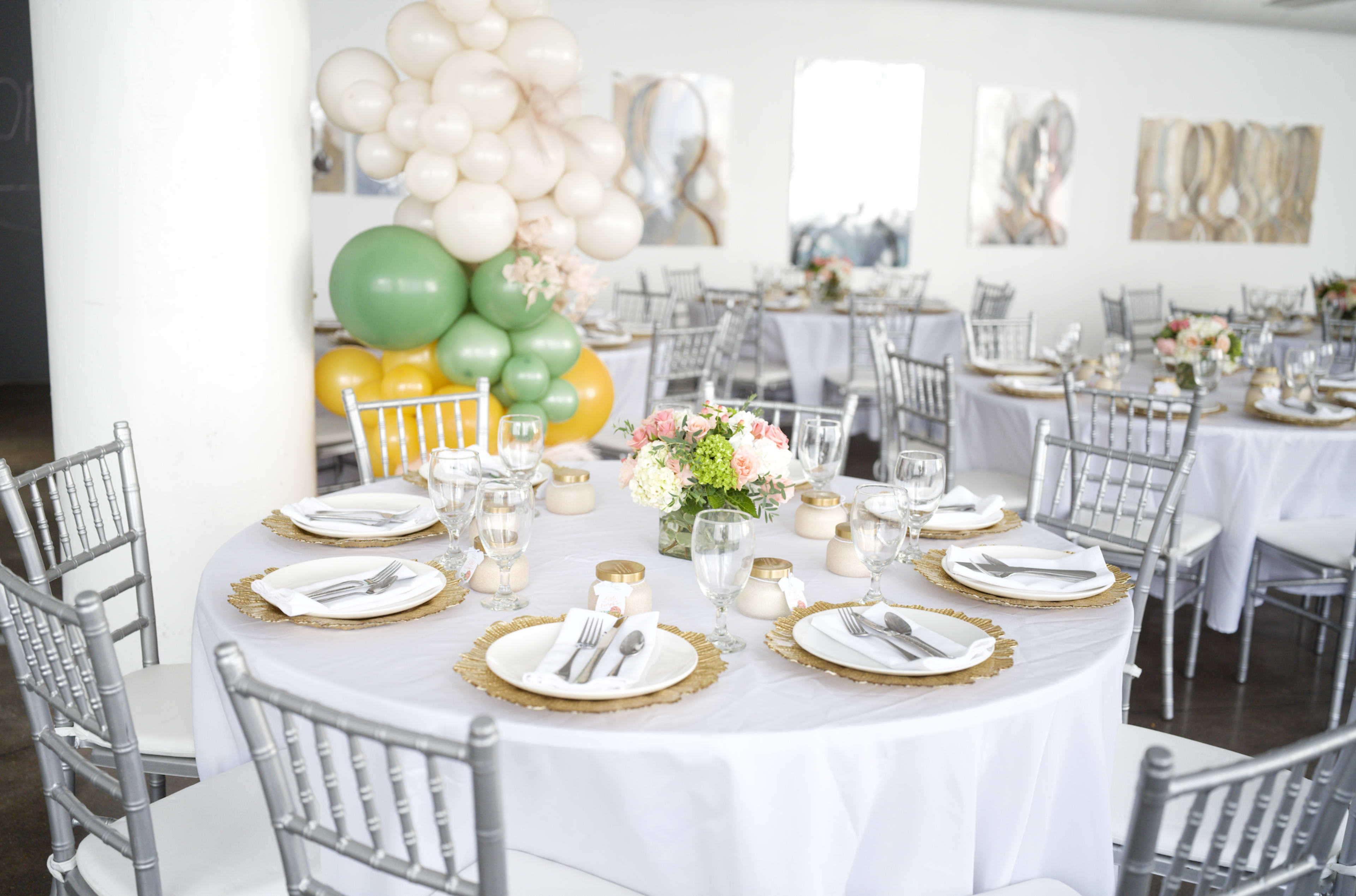 A banquet setting features elegantly arranged tables with white linens, gold-rimmed plates, and decorative floral centerpieces, alongside a backdrop of colorful balloon arrangements.