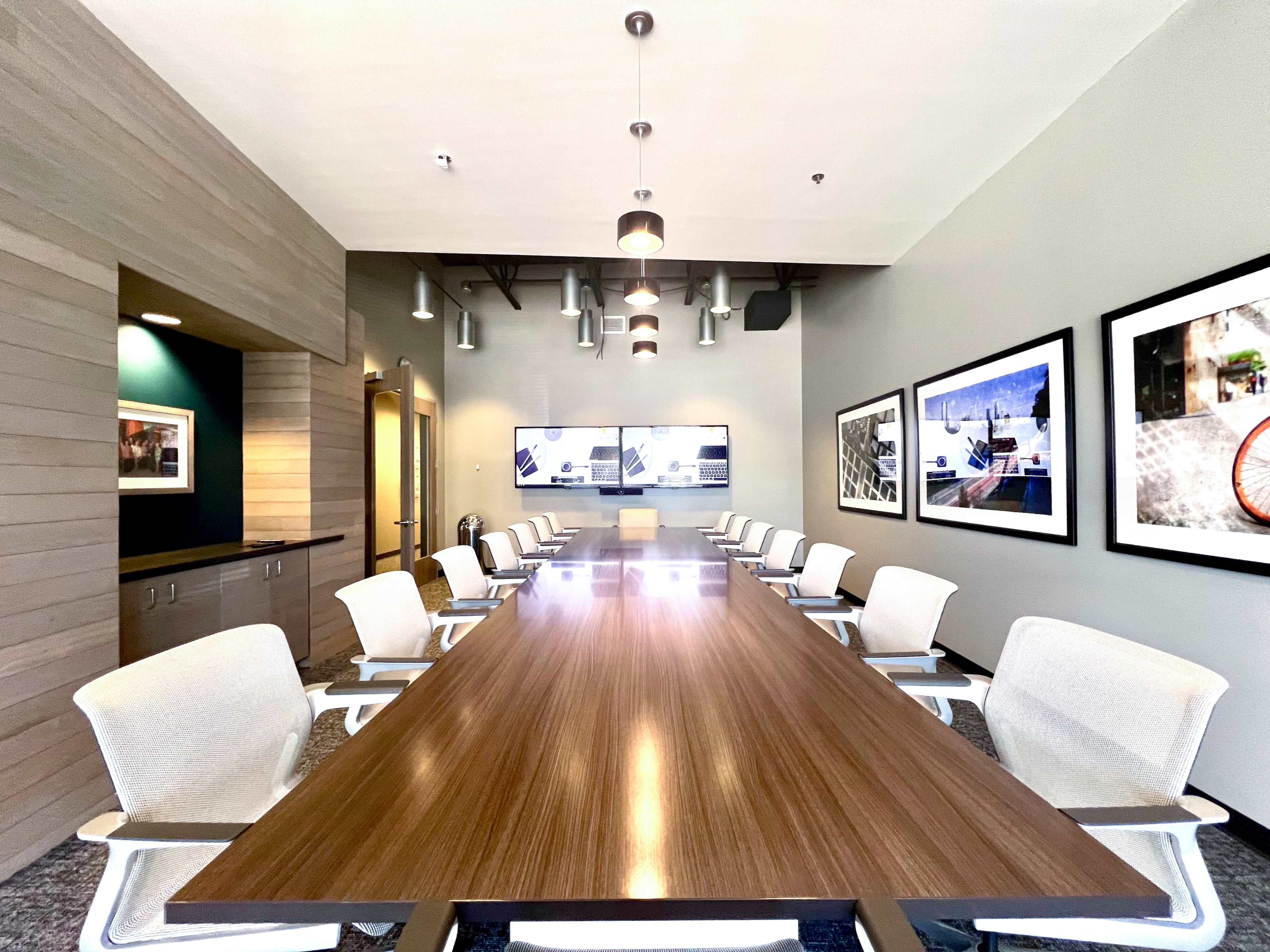 The image shows a modern conference room with a long wooden table and several white chairs, featuring wall-mounted screens and framed photographs on the walls.