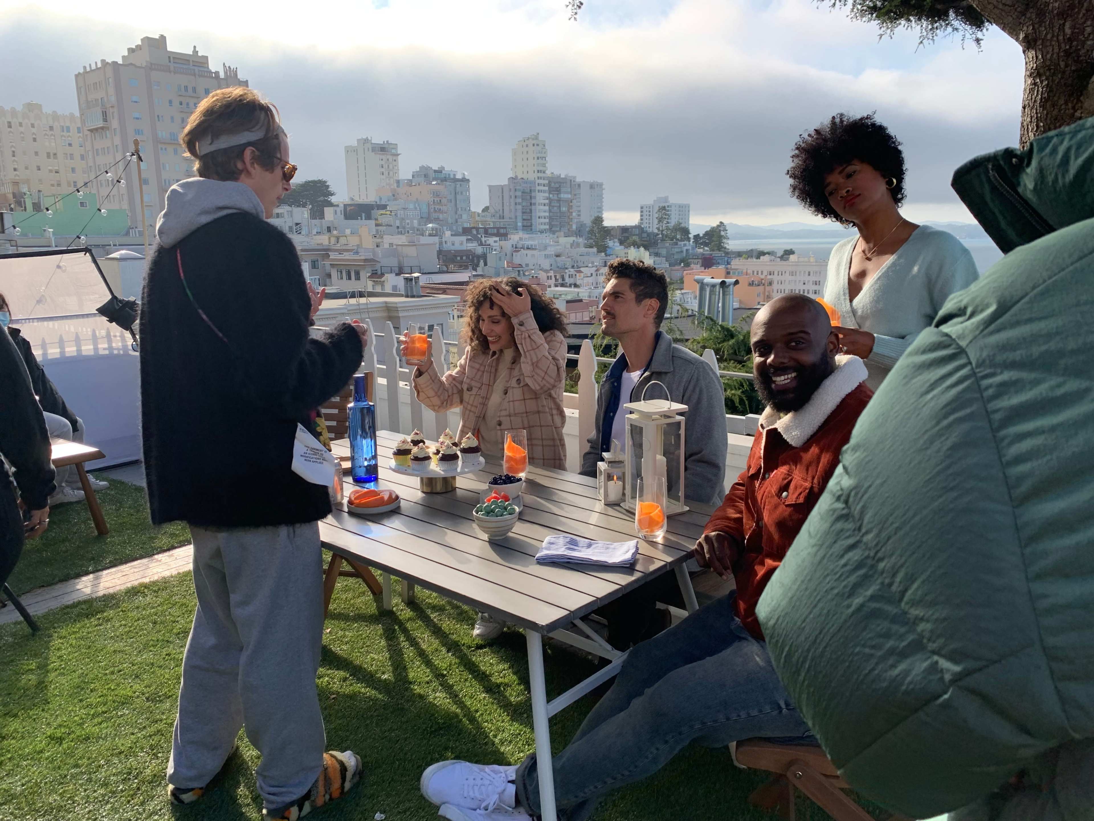 A group of five people is gathered around a table on a patio with a view of a city skyline, enjoying drinks and conversation.