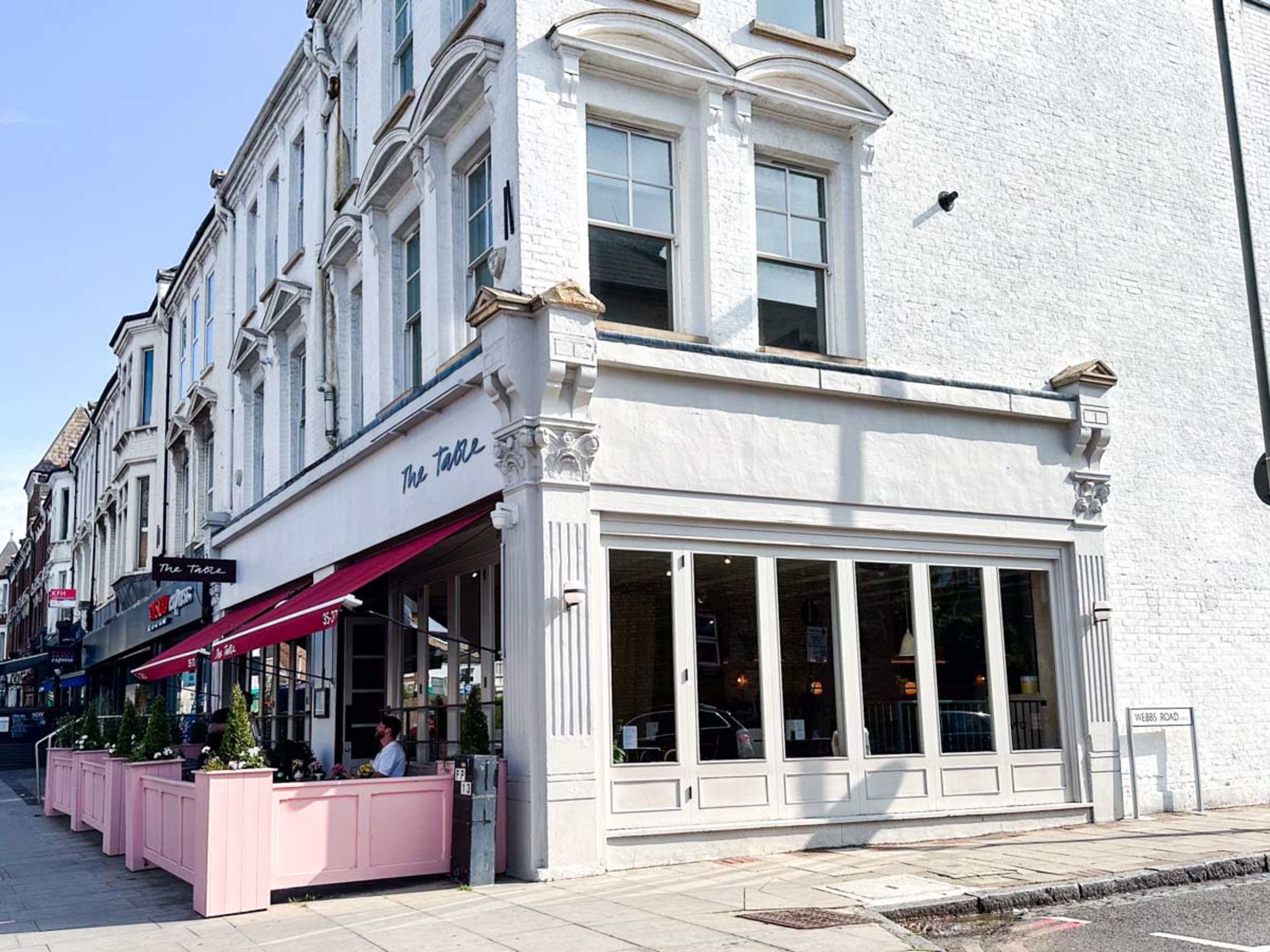 A restaurant with a white exterior and large windows, featuring outdoor seating and decorative plants along the sidewalk.