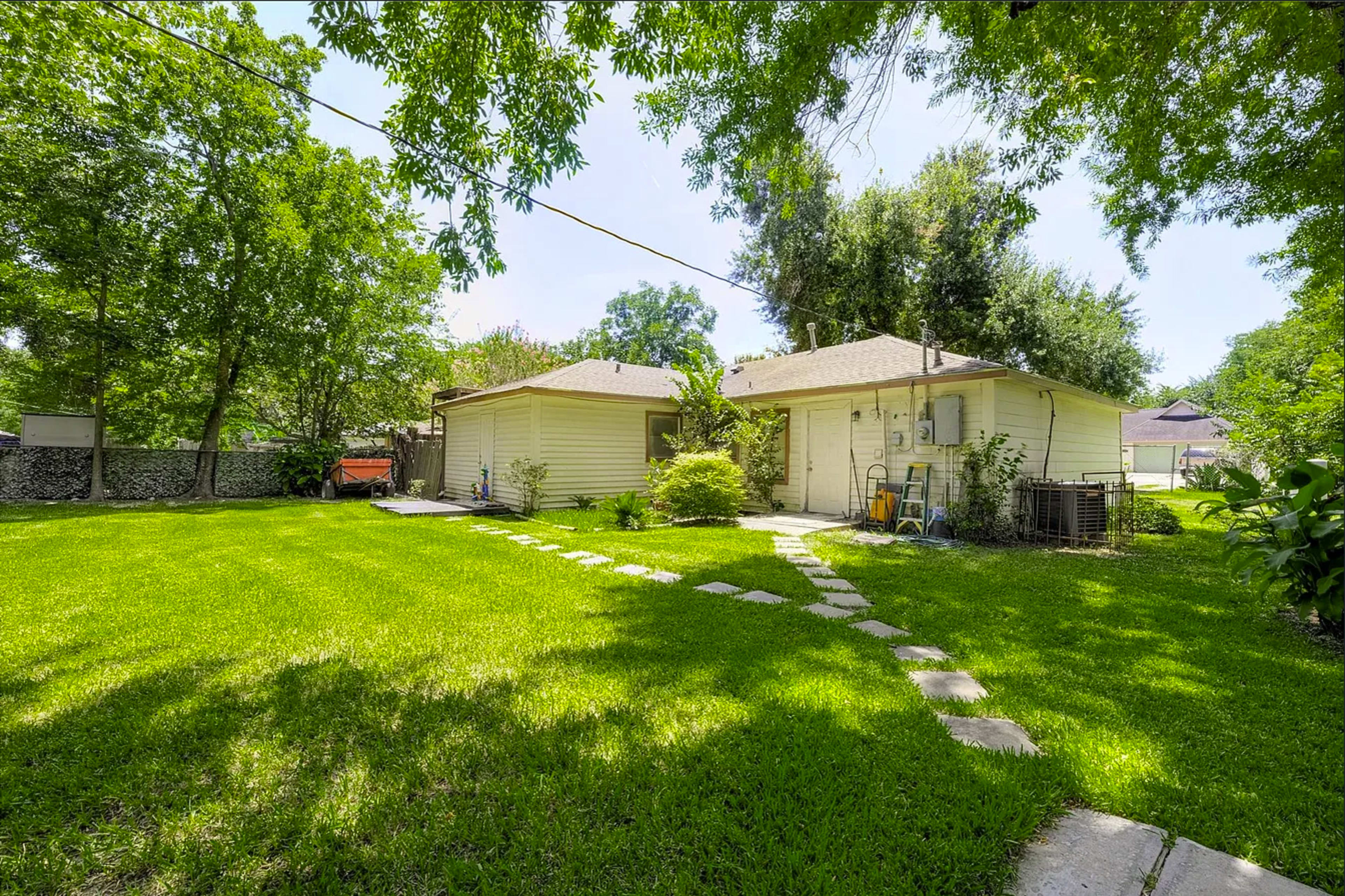 The image shows a grassy yard with a pathway made of stones leading to a yellow house surrounded by trees.