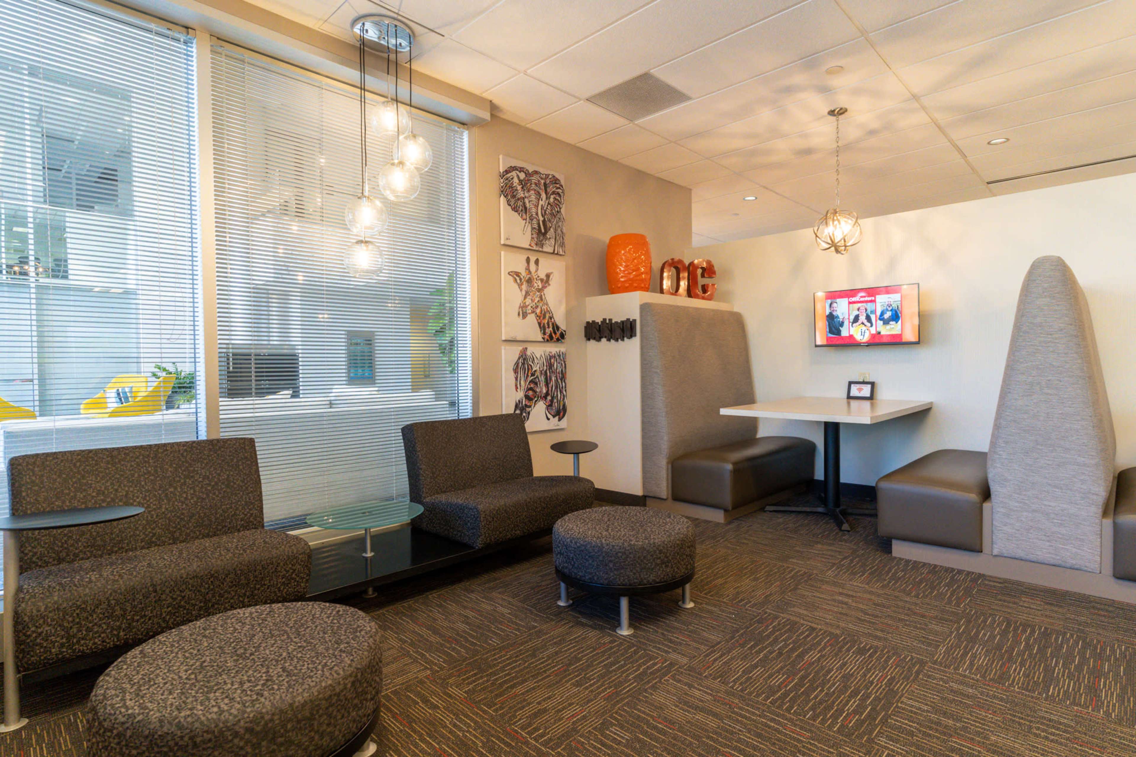 The image shows a modern lounge area with circular seating, a small table, and a television on the wall, surrounded by glass walls that provide a view of an office space.