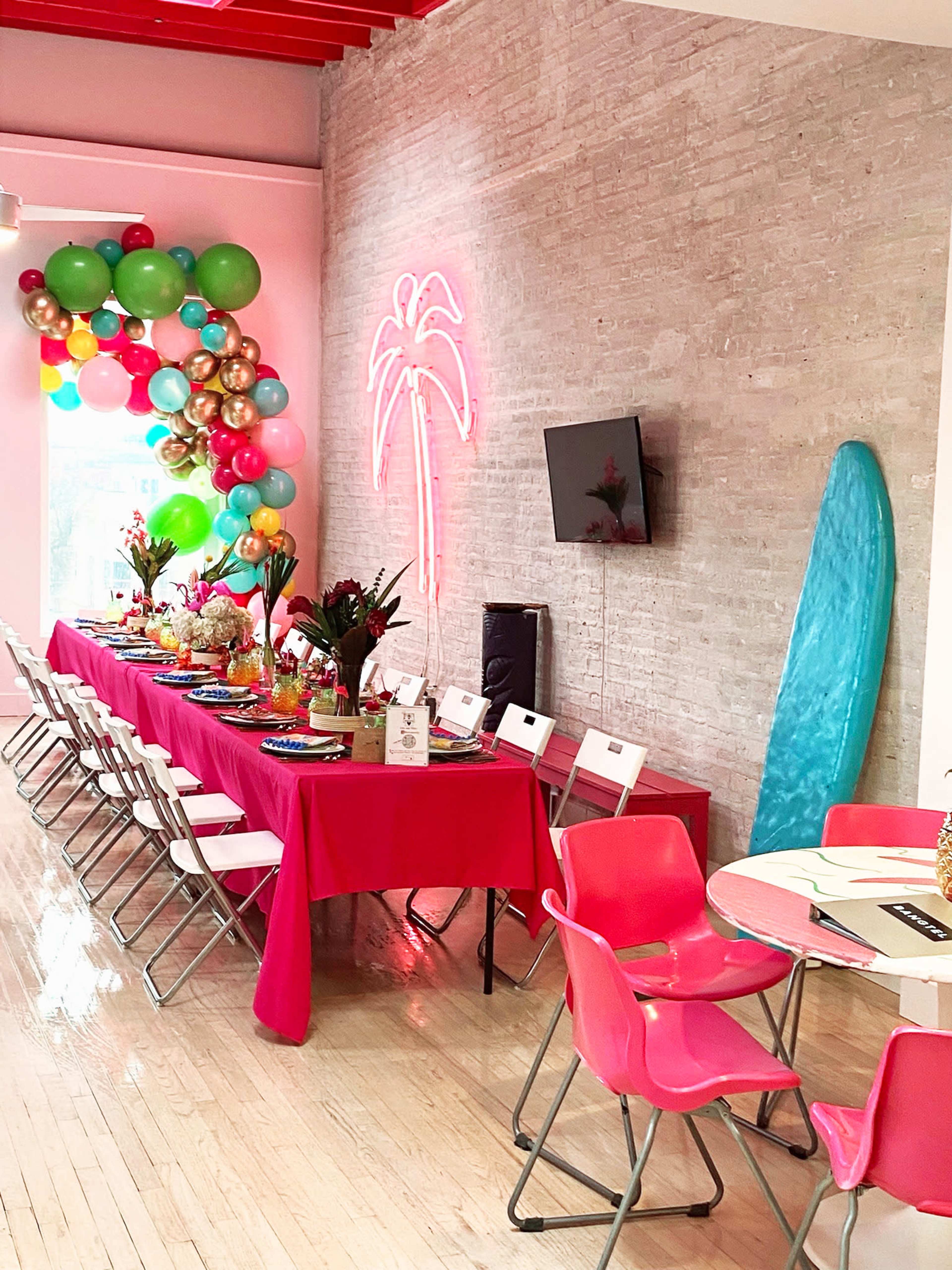 A vibrant party setting features a long table adorned with colorful decorations, surrounded by chairs, with balloons and a neon palm tree sign accentuating the room.