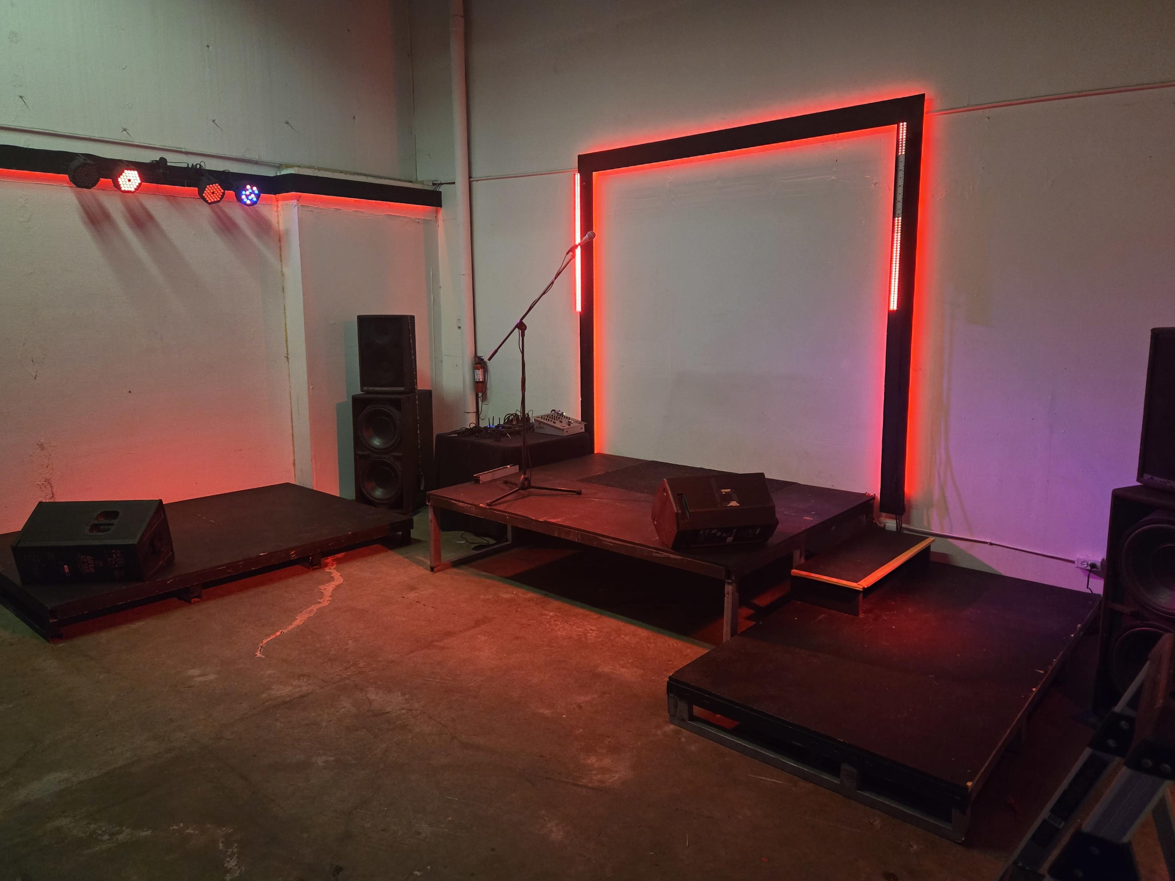 The image shows a small stage setup with two elevated platforms, a microphone mounted on a stand, and colorful lighting along the edges, all positioned in an industrial space.