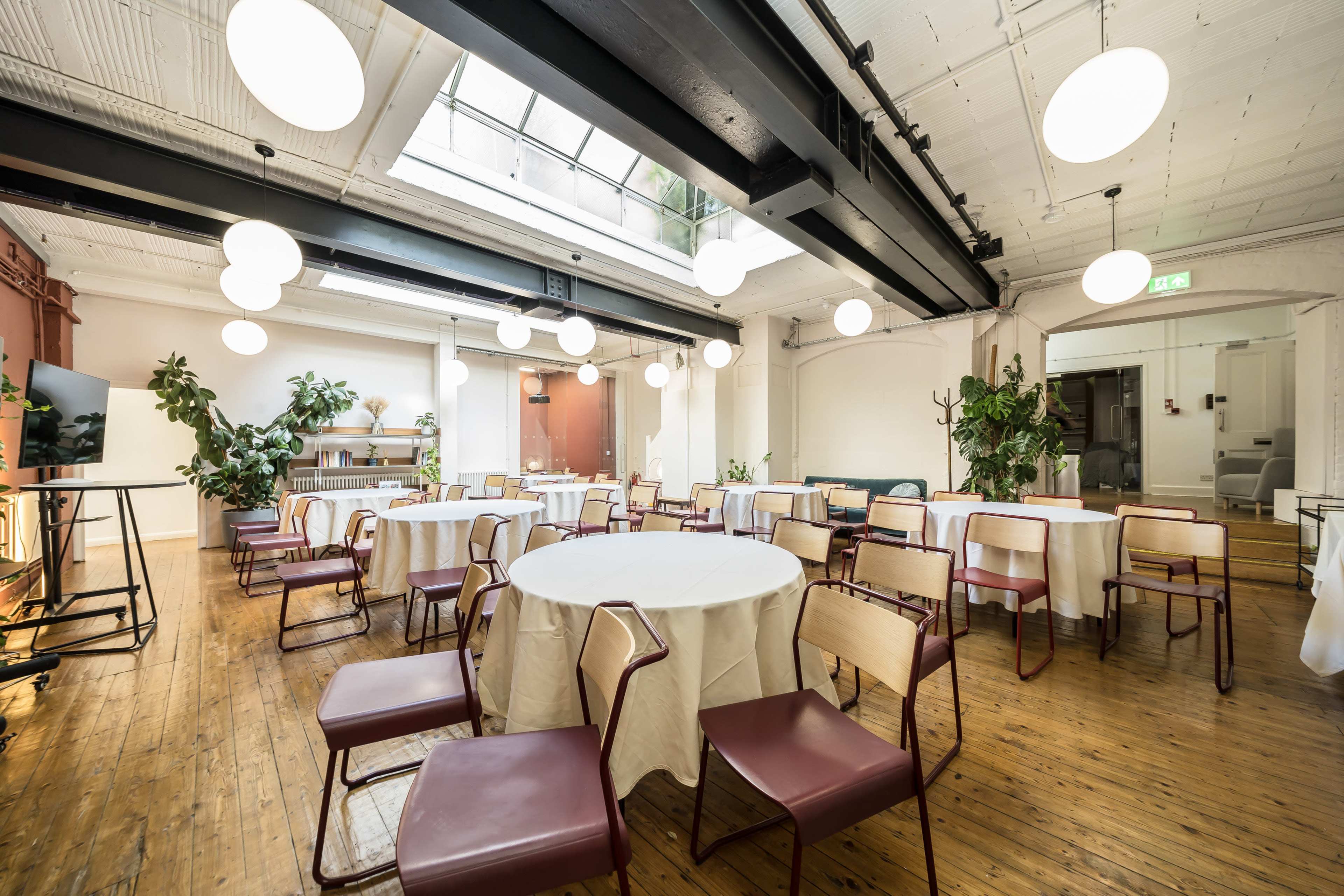 The image shows a well-lit indoor venue featuring several round tables covered with white tablecloths and surrounded by chairs, with plants and dim pendant lights in the background.