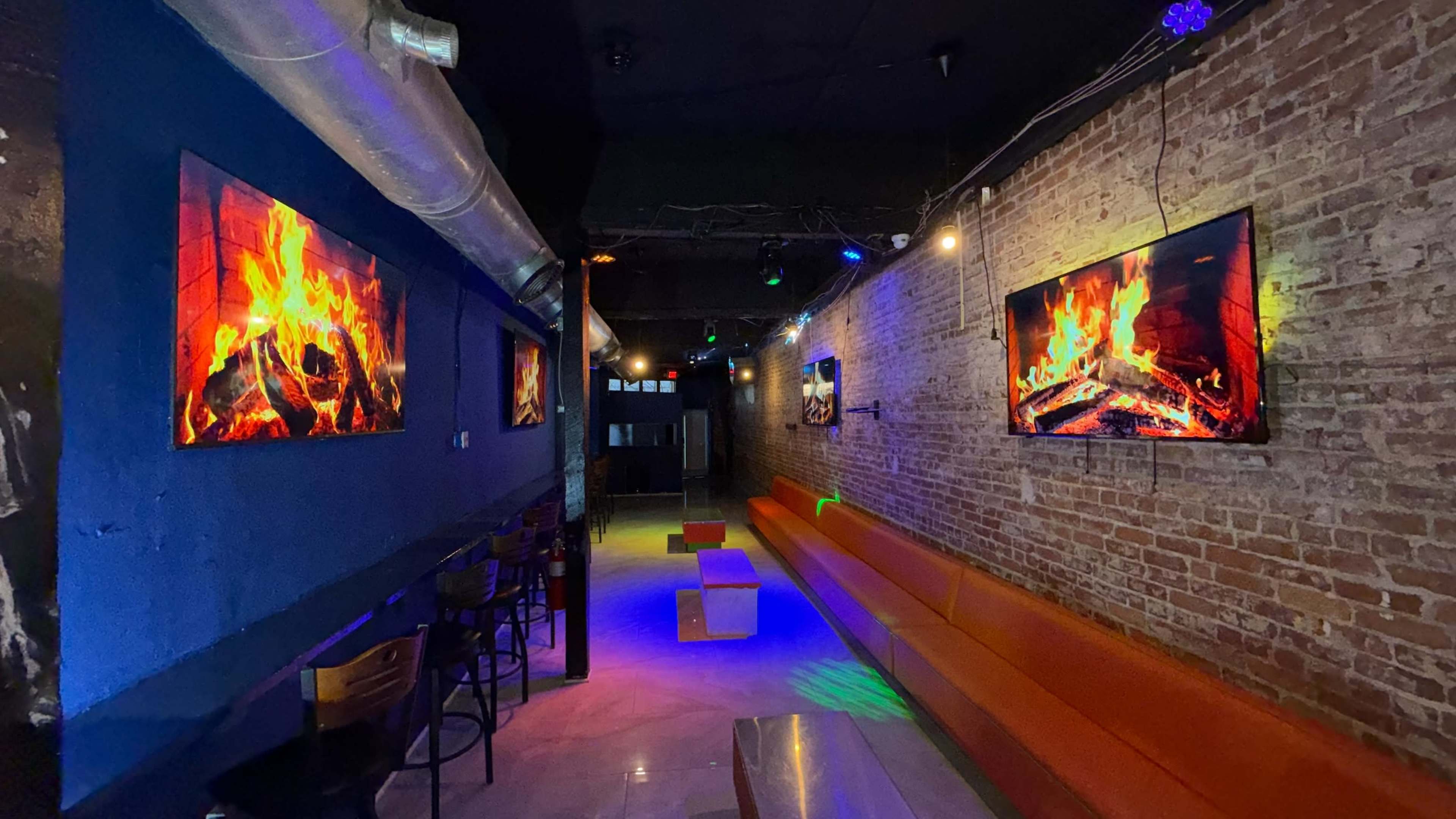The image shows a dimly lit lounge with brick walls, featuring large screens displaying fire visuals and a long orange seating area.