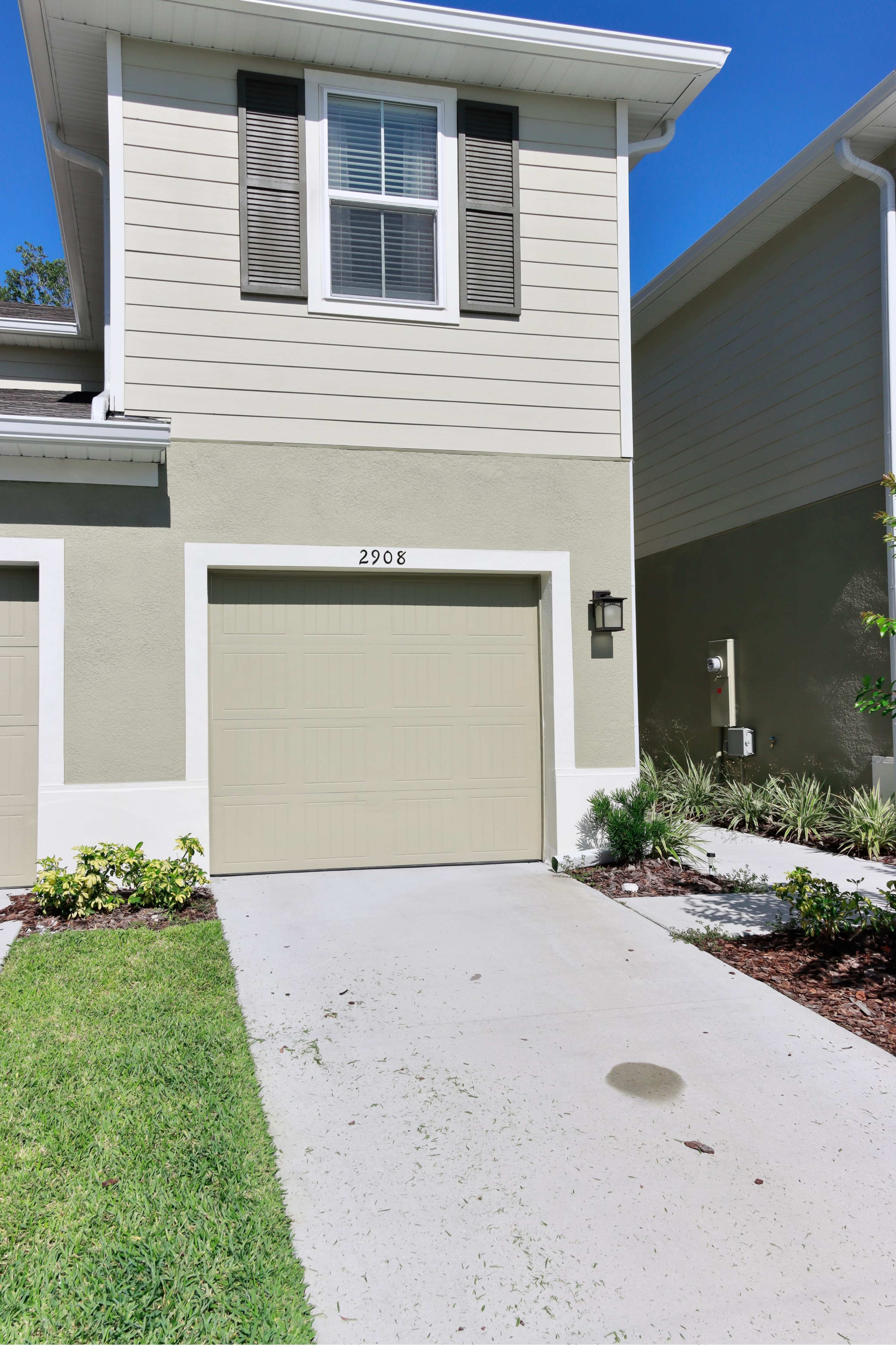 A two-story residential building with a garage door and landscaped front area, featuring a small driveway and green plants.
