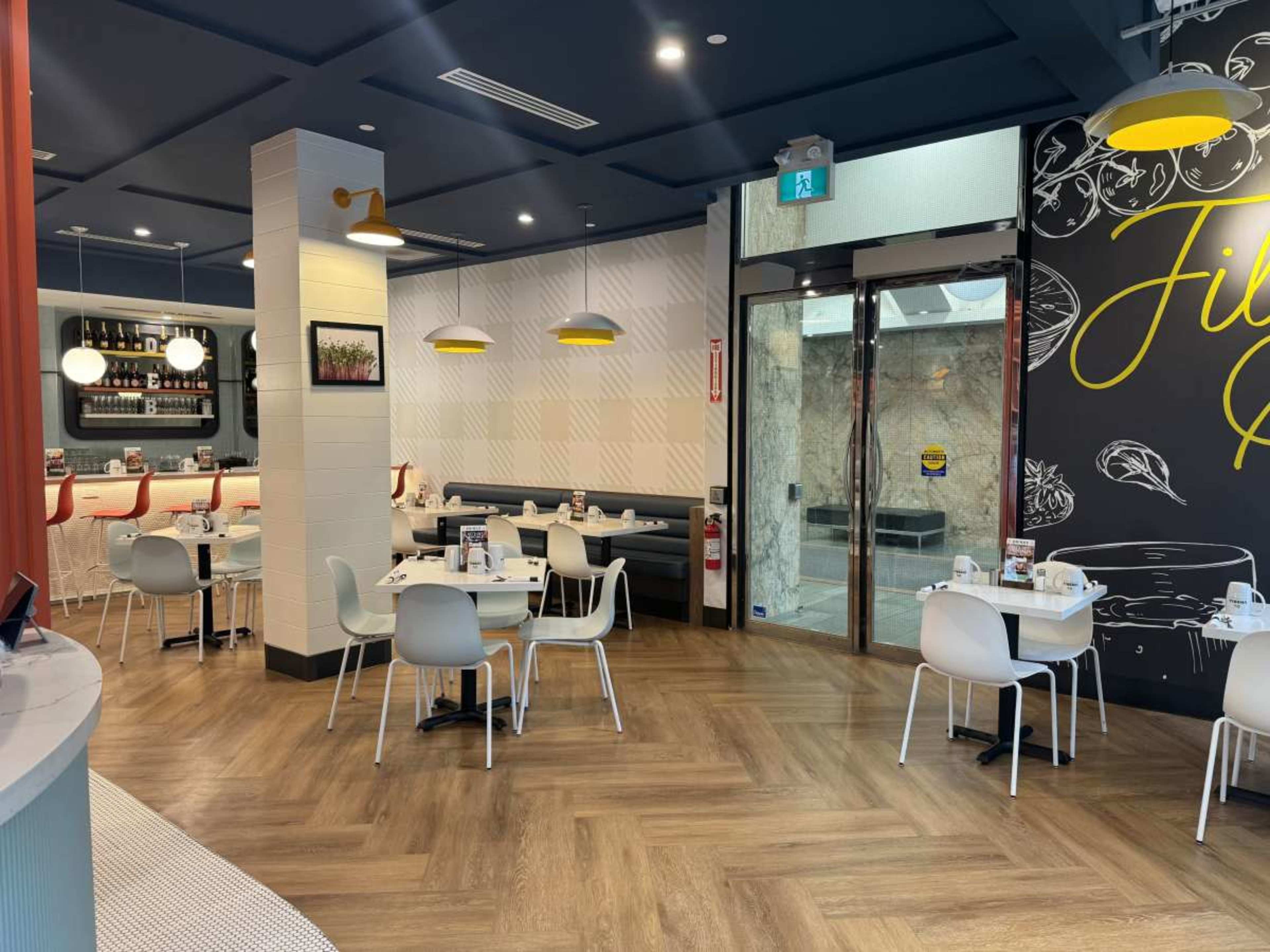 The image shows a modern restaurant interior with white tables and chairs, a bar area with wine bottles, and large windows illuminated by yellow pendant lights.