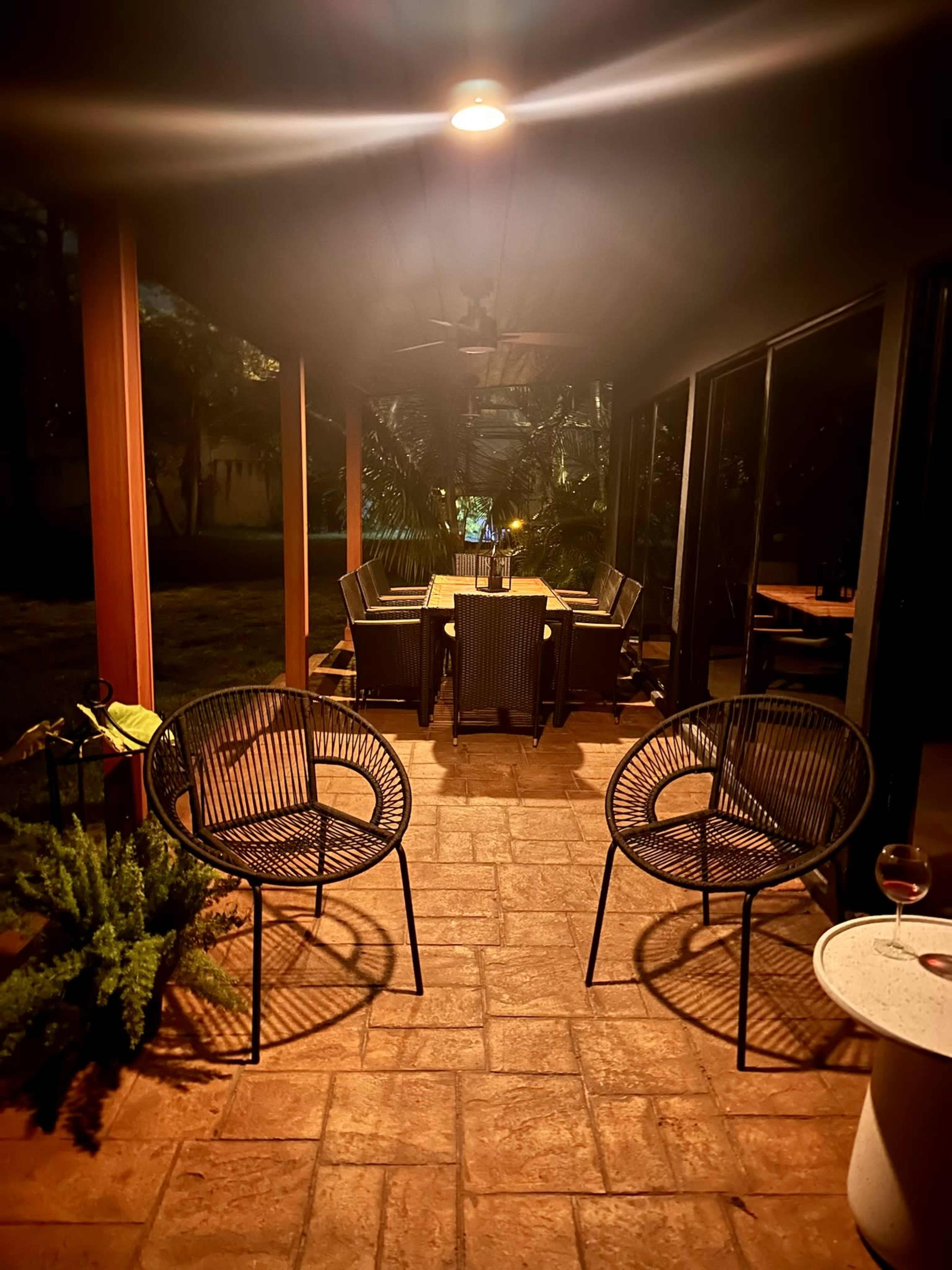 A well-lit patio features a dining table surrounded by several chairs, with two round chairs positioned nearby and a glass of wine on a small table.