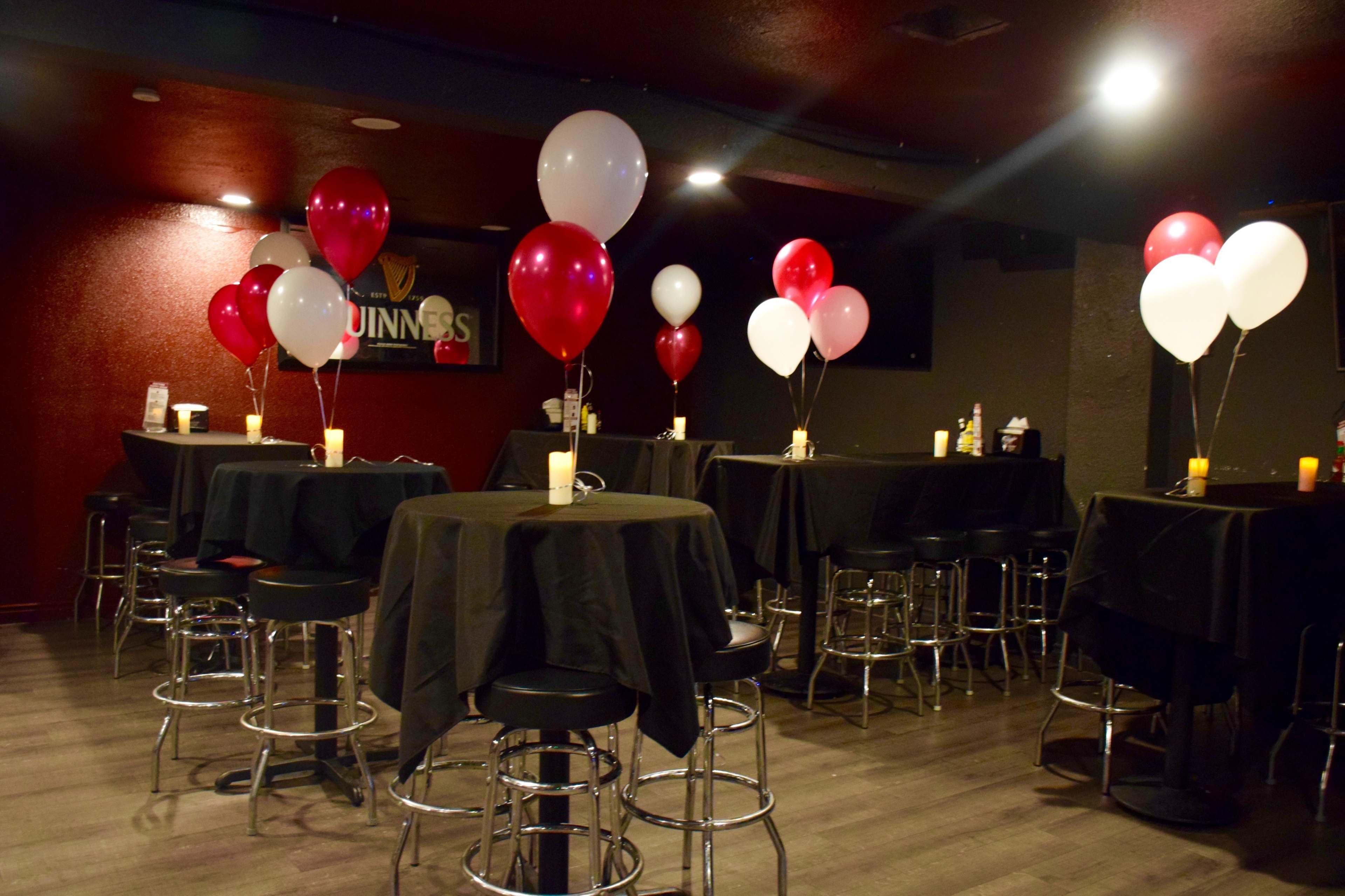 A dimly lit party venue with black tablecloths, high-top tables adorned with red and white balloons, and flickering candlelight on each table.