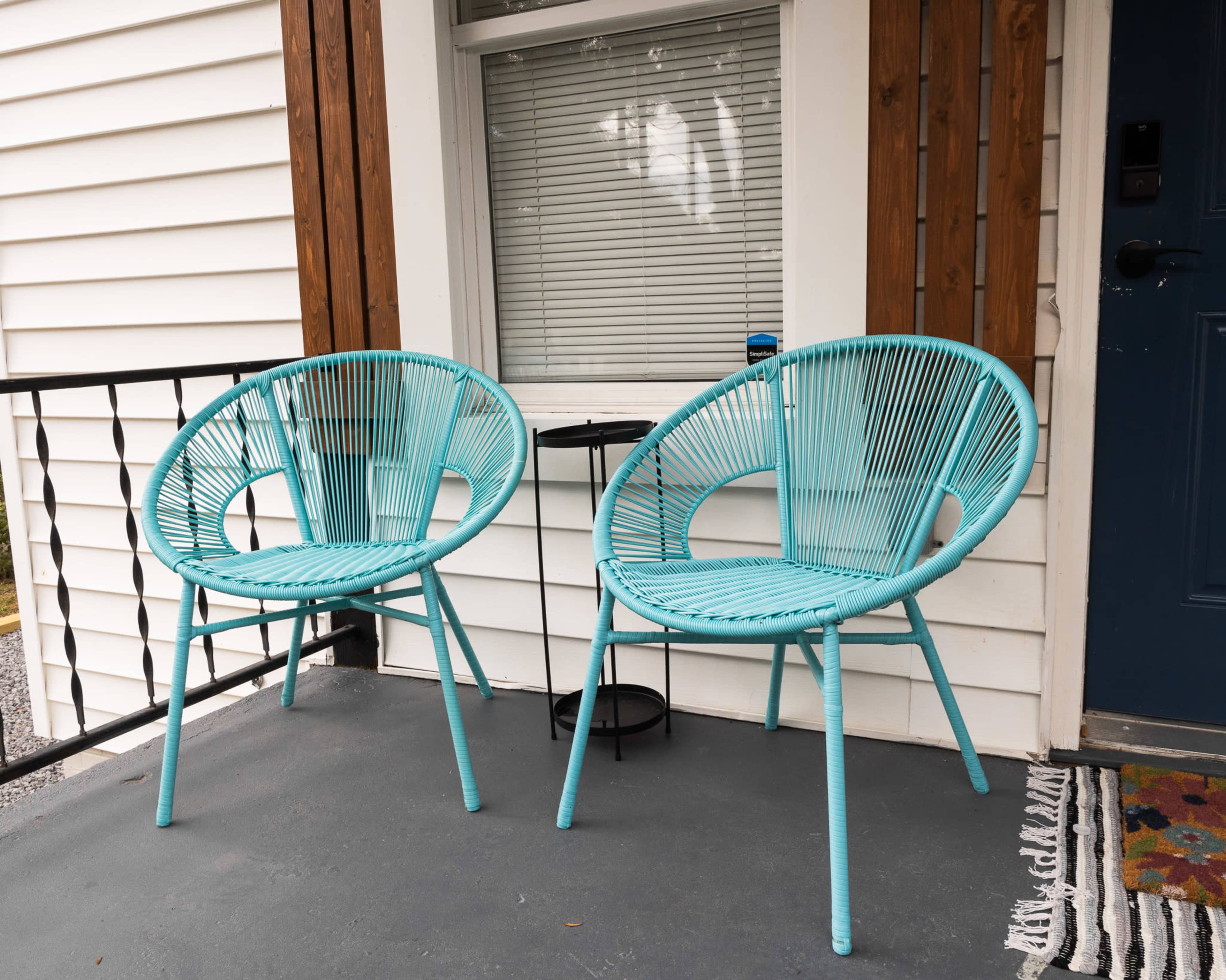 Two turquoise chairs are positioned on a porch next to a small round table, with a gray floor and white siding in the background.