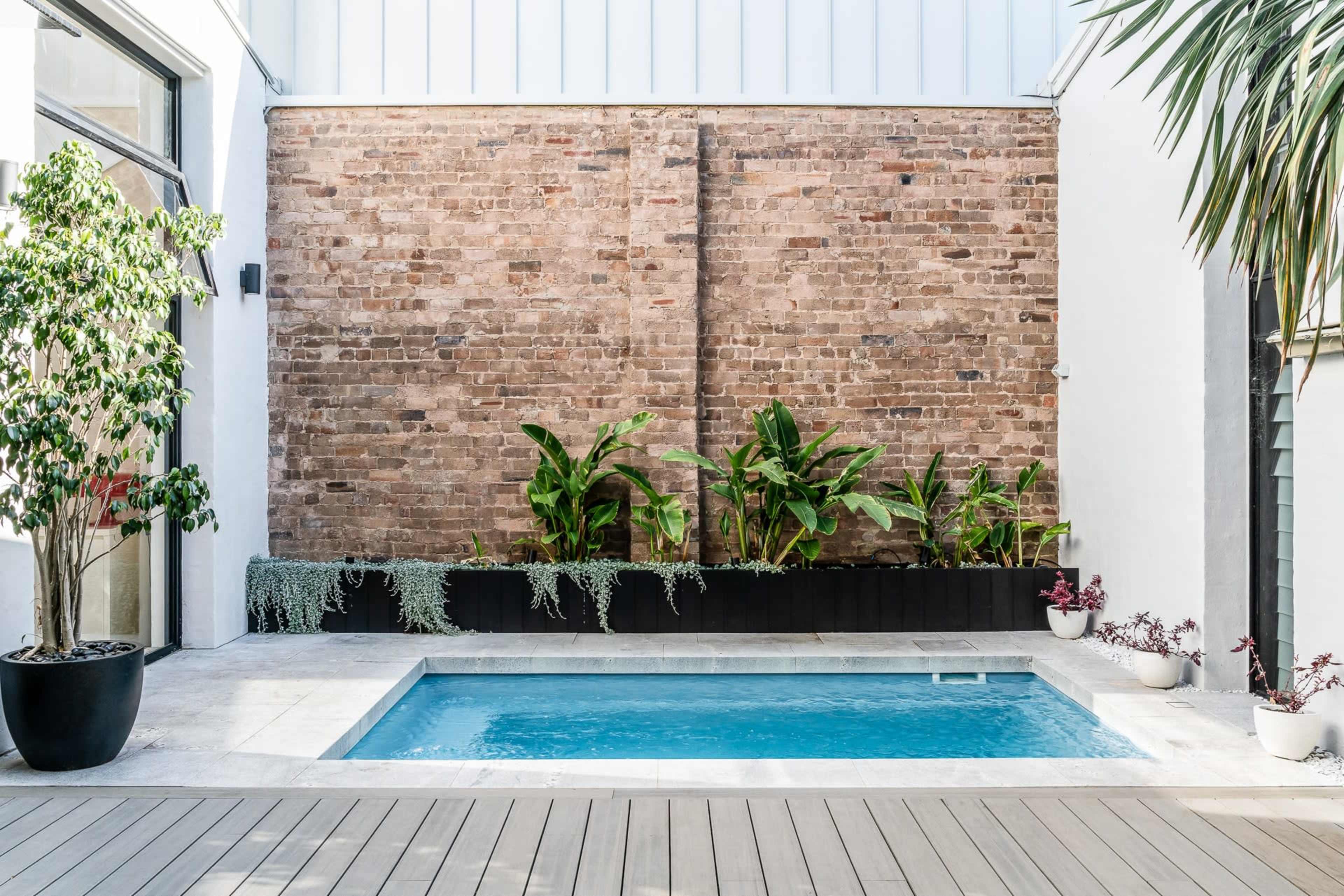 A small swimming pool set against a brick wall, surrounded by potted plants and a wooden deck.