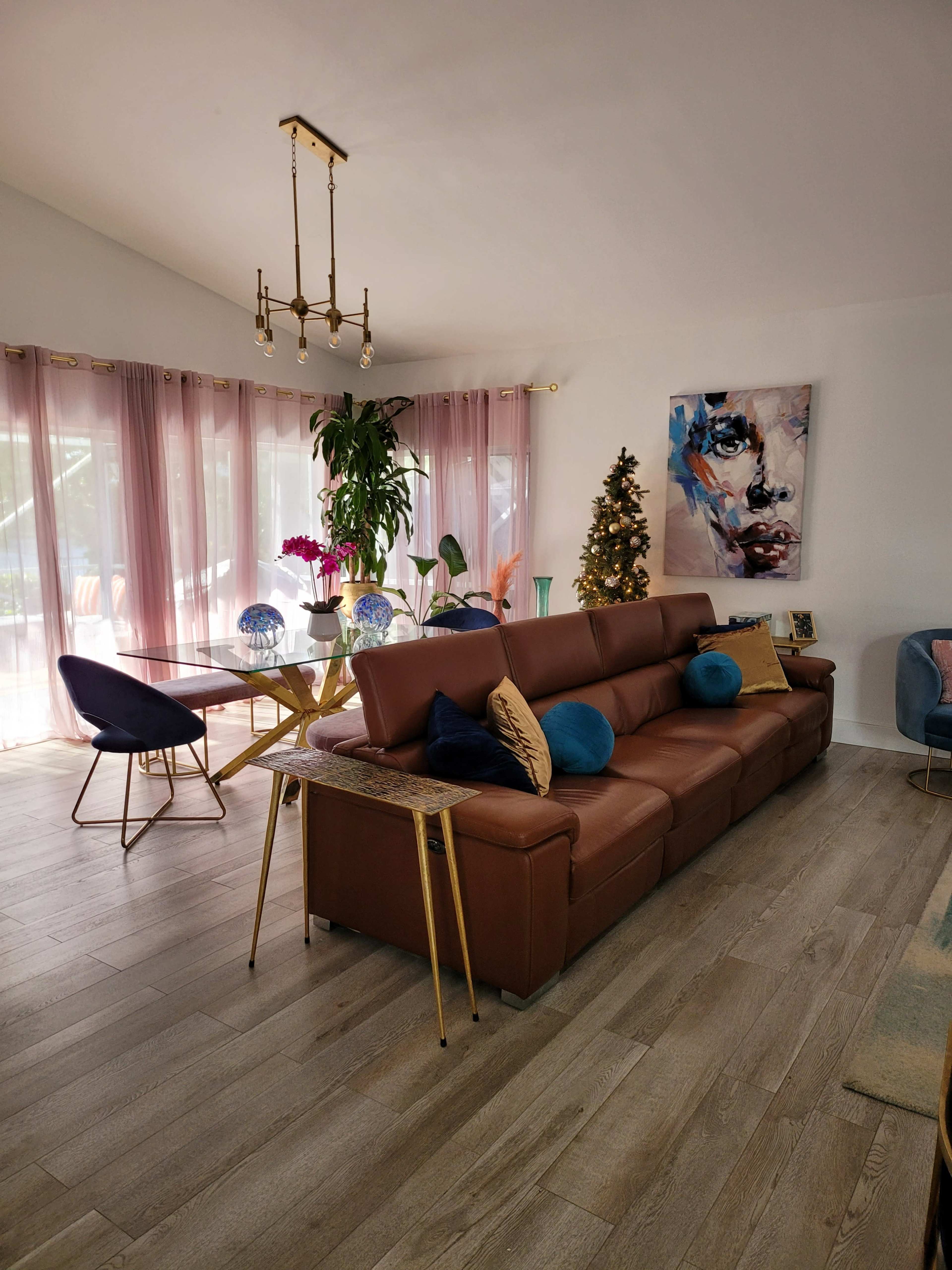 A modern living room features a brown leather sofa with colorful throw pillows, a glass dining table, and a Christmas tree in the corner.
