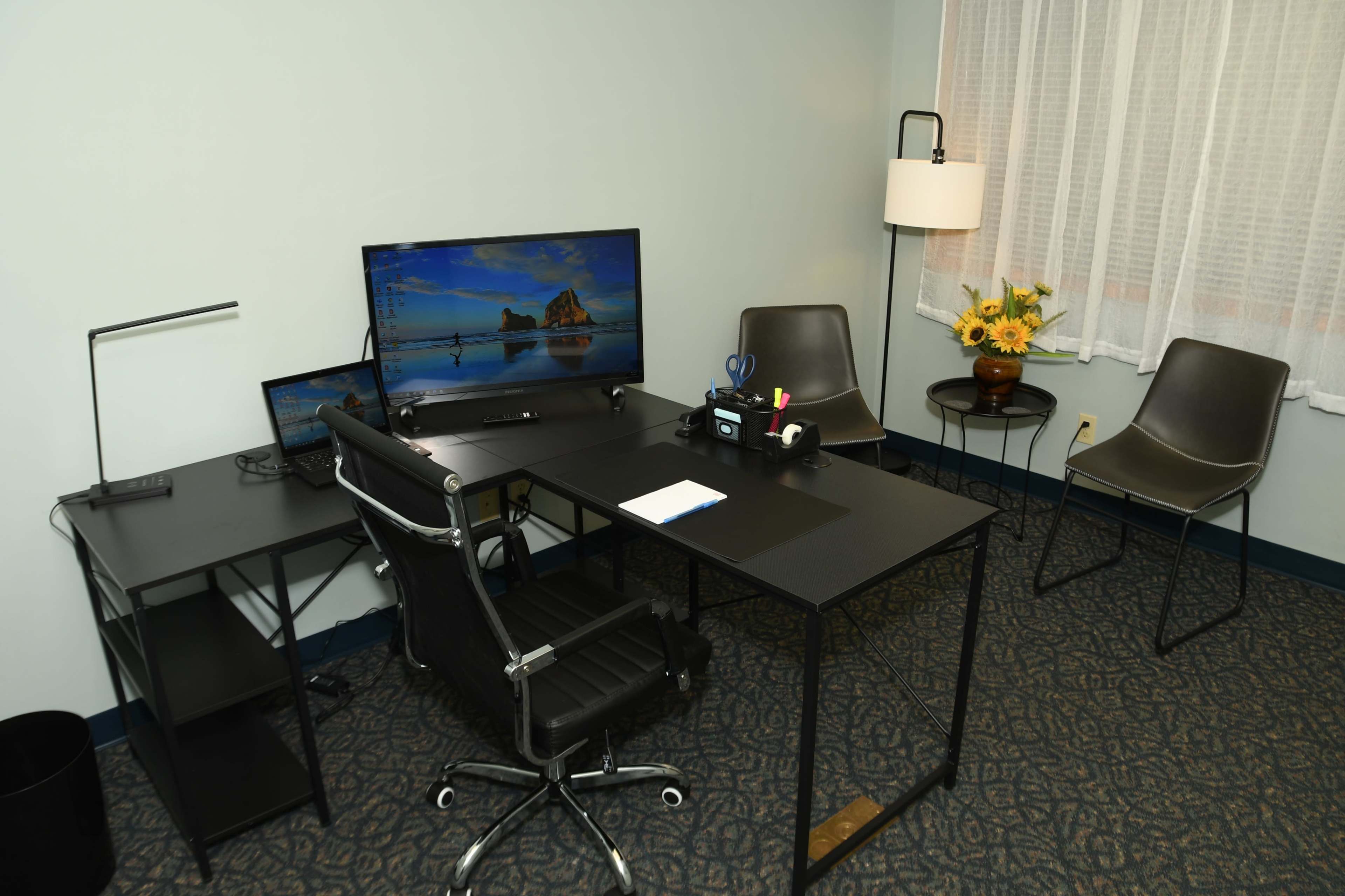 The image shows a modern office space equipped with a desk, two monitors, a black office chair, and a small seating area with two chairs and a lamp.