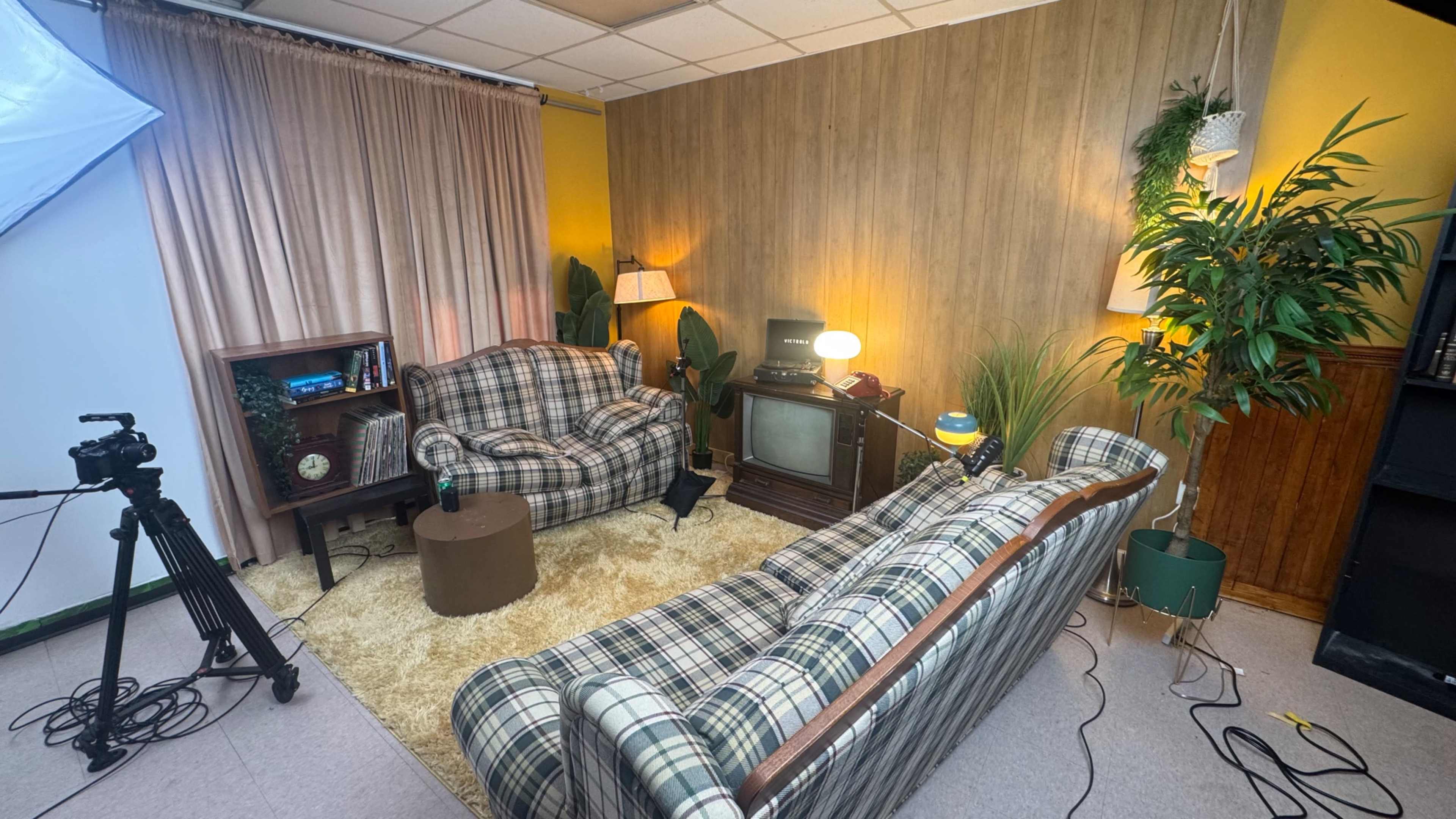 The image shows a cozy living room setup with two plaid sofas, a round coffee table, an old television, and plants against a backdrop of wood paneling and yellow walls.