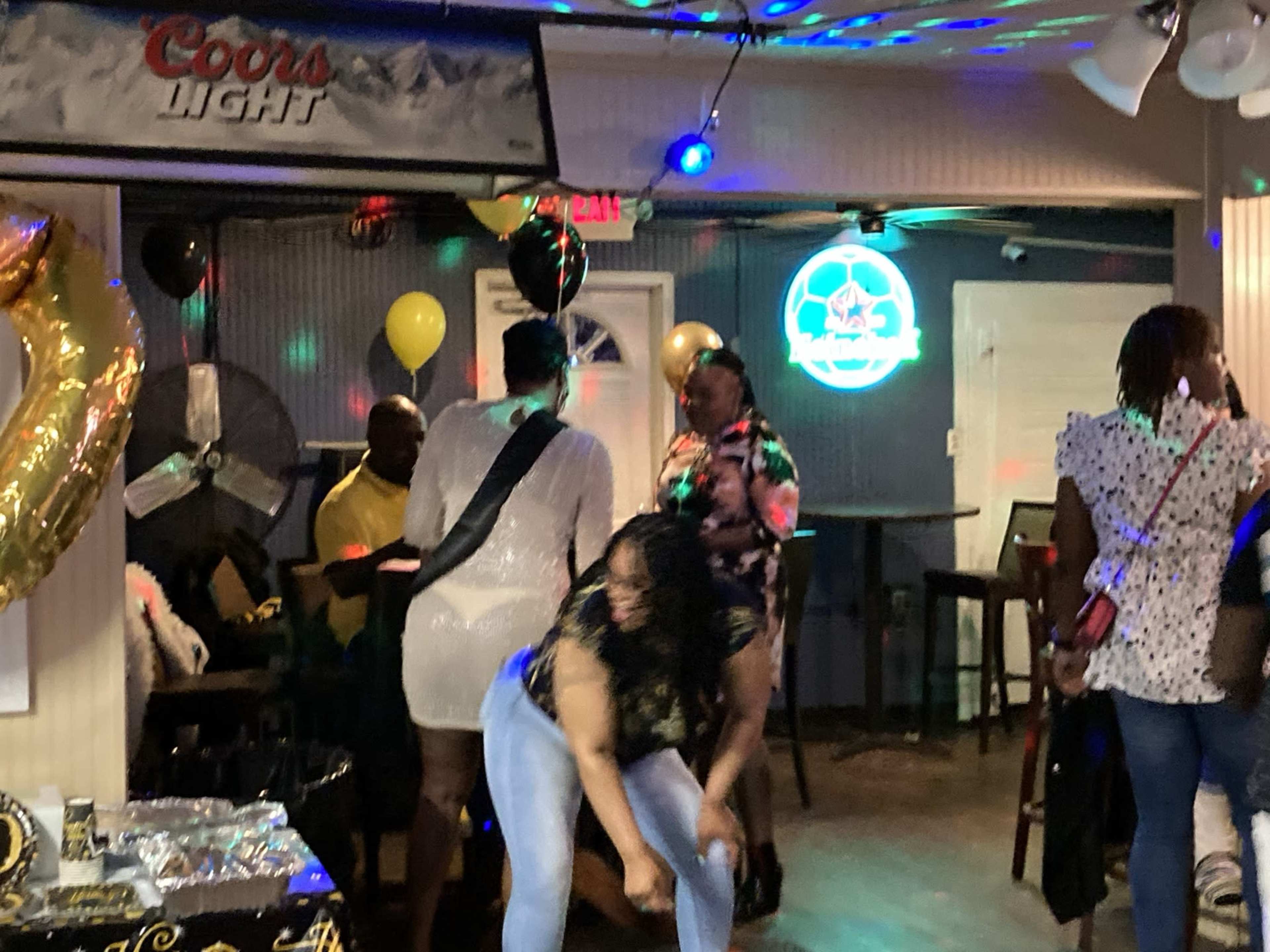 A group of people socialize and dance in a brightly lit bar decorated with balloons and banners.