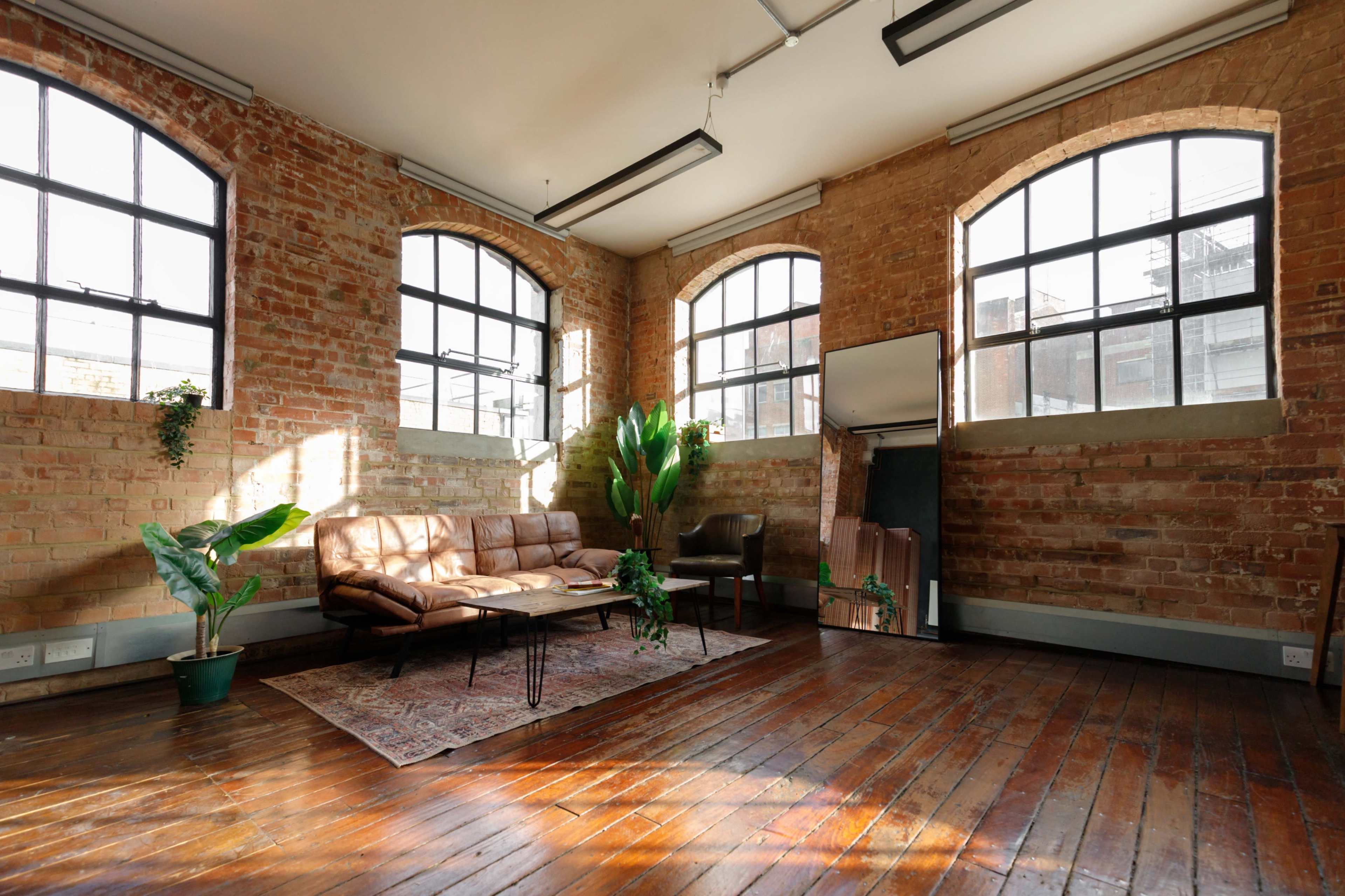 The image shows a spacious room with exposed brick walls, large windows, a brown leather sofa, a coffee table, a green plant, and hardwood flooring.