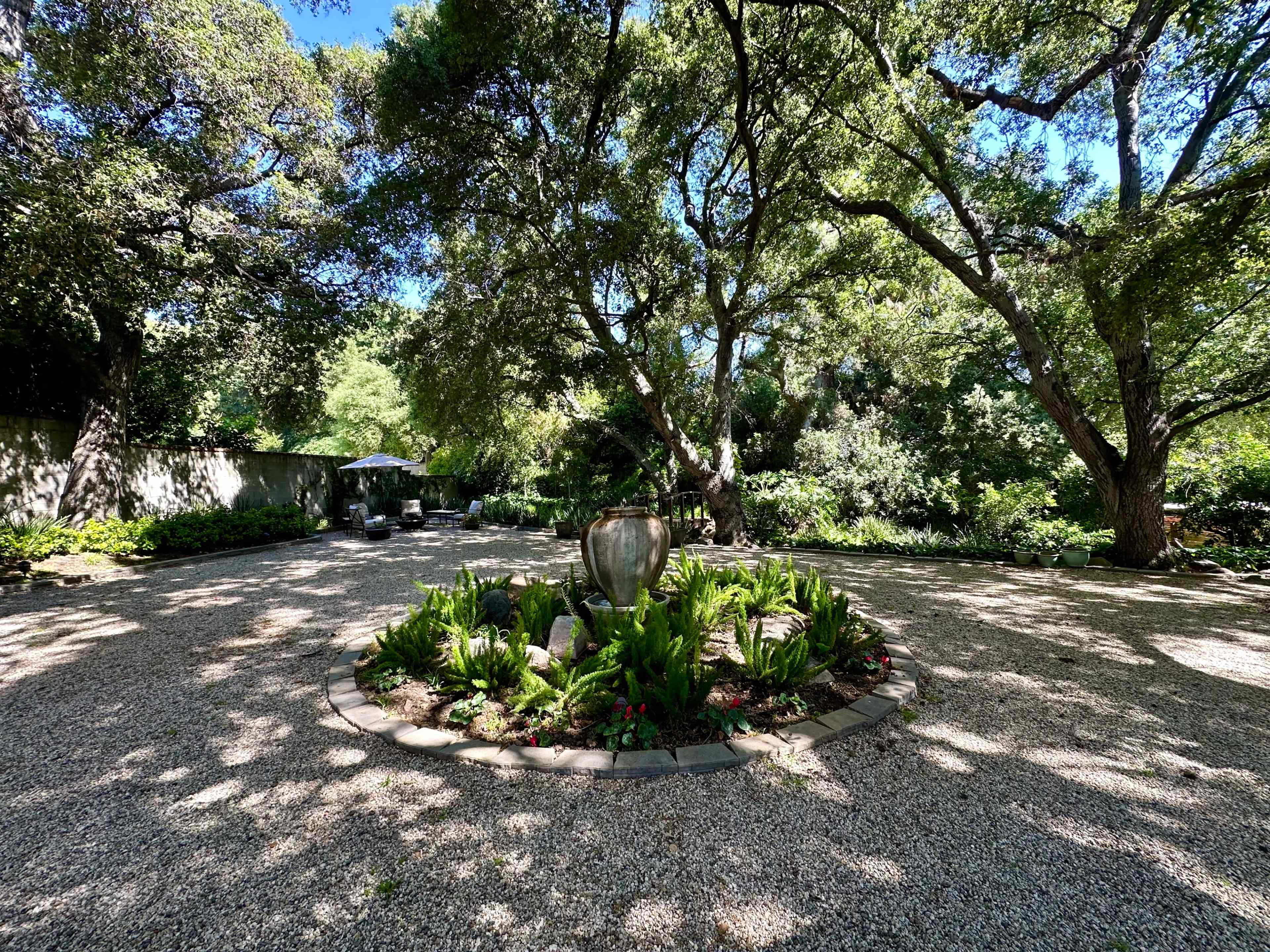 A circular garden space features a stone fountain surrounded by lush greenery and gravel pathways under a canopy of large trees.