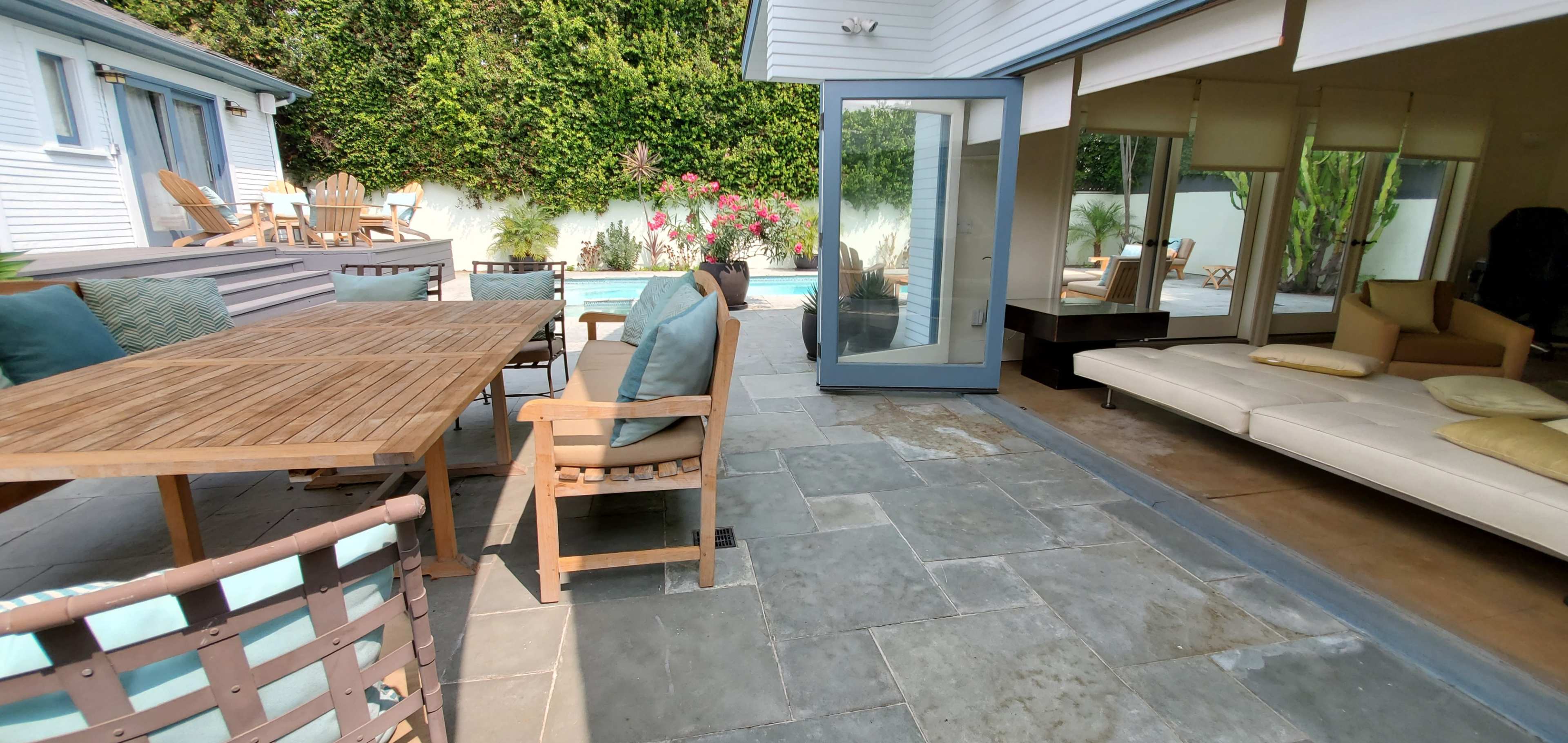 A patio area with a wooden dining table and chairs beside a pool, adjacent to a modern interior space with large glass doors.