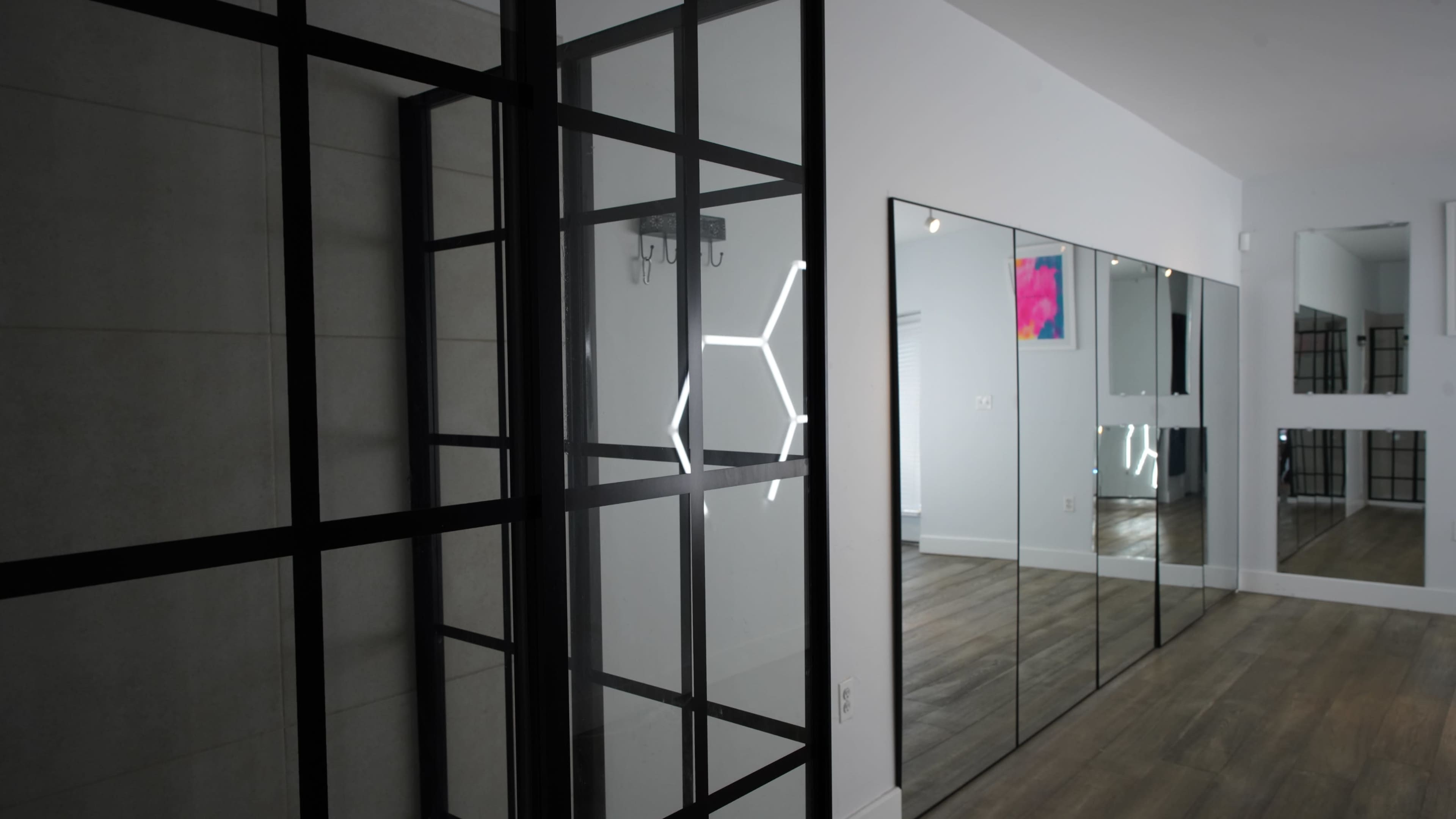 The image shows a hallway featuring mirrored walls and a black-framed glass partition illuminated by a geometric light fixture.