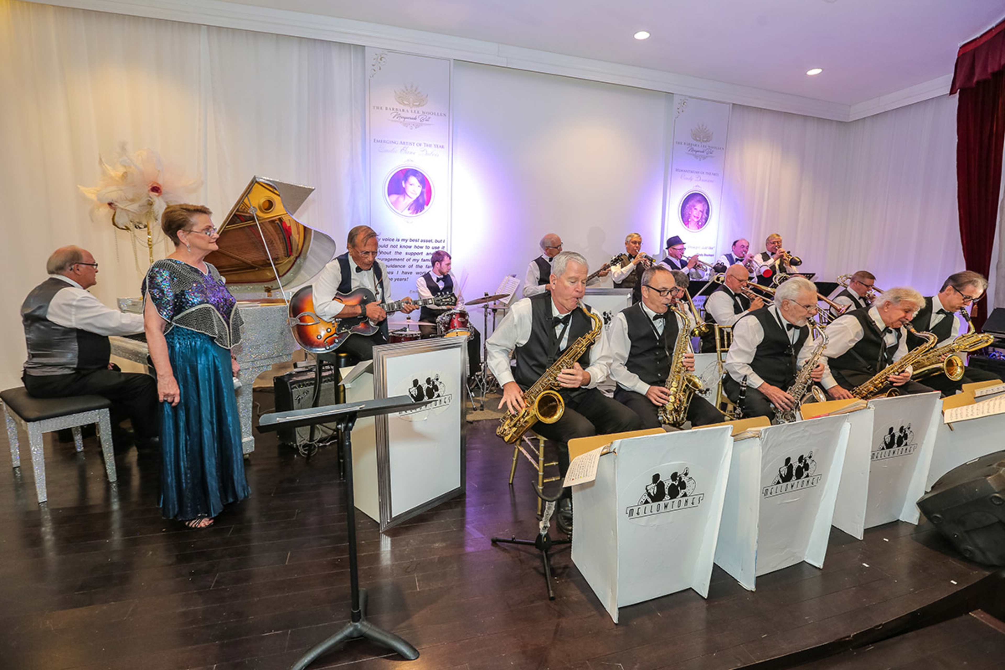 A large orchestra with musicians in formal attire performs on stage, accompanied by a female vocalist in a blue dress.