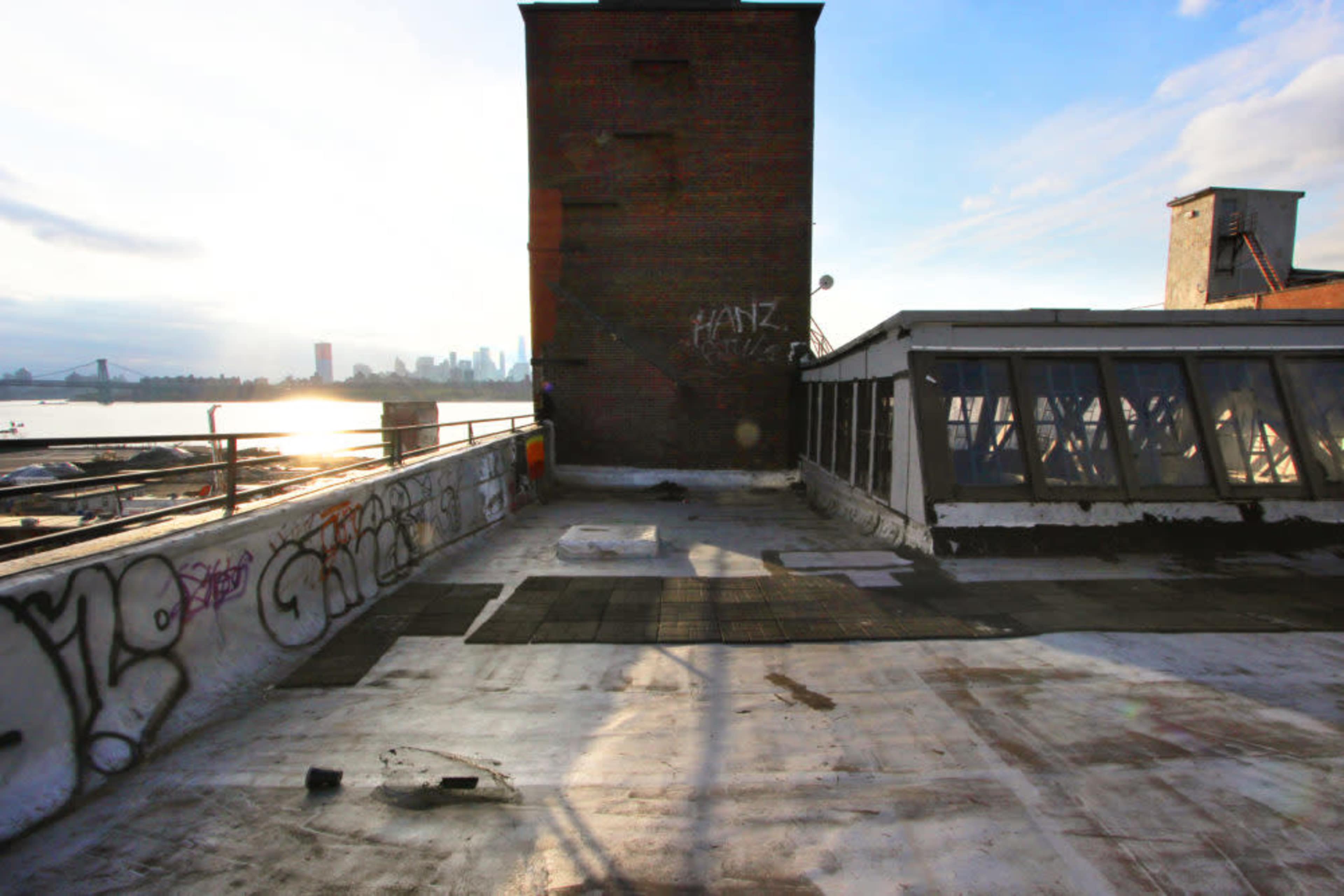 The image shows a rooftop with a brick structure and graffiti, overlooking a river and a city skyline in the background.