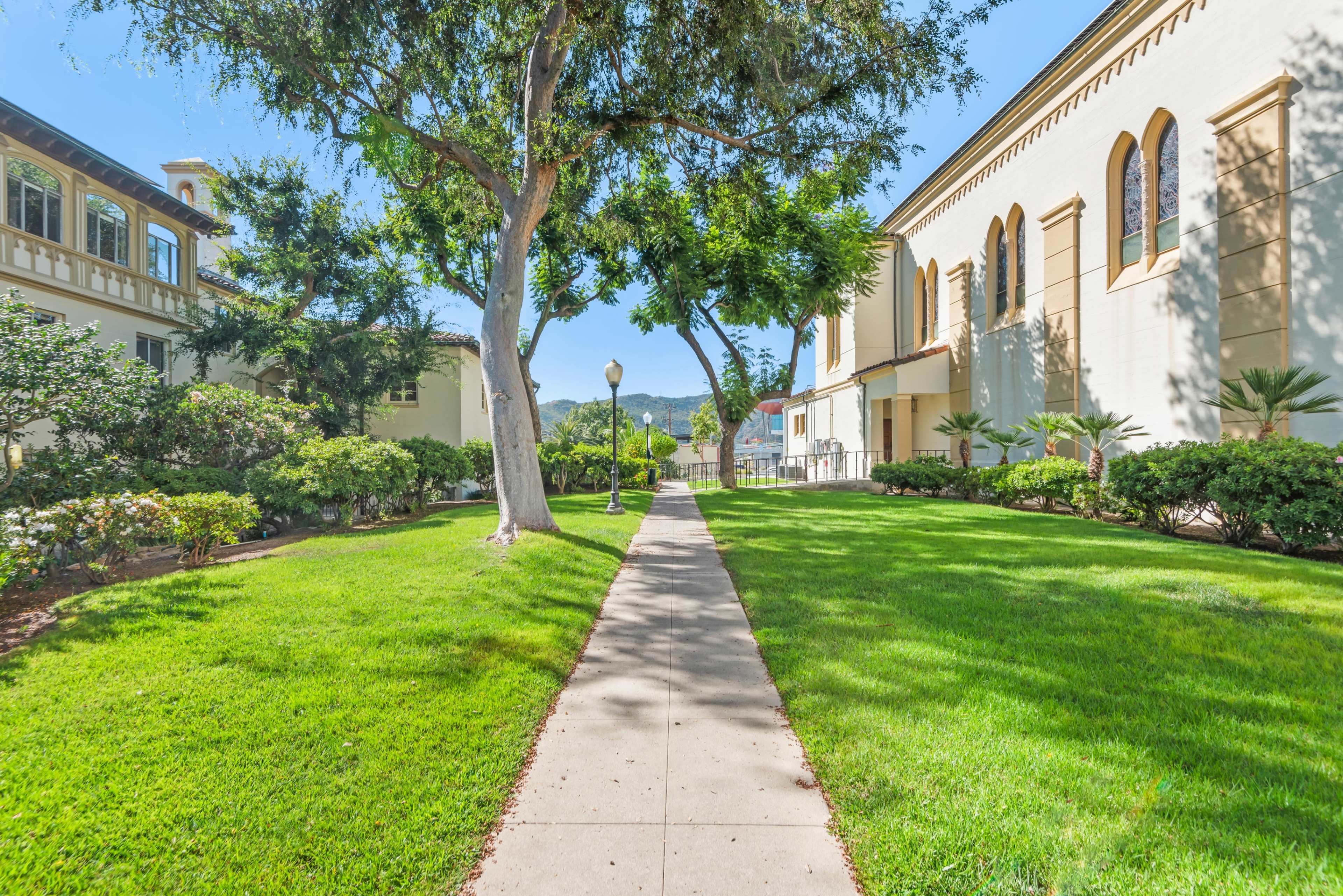 A paved walkway lined with grass and trees leads past buildings with architectural details in a landscaped area.