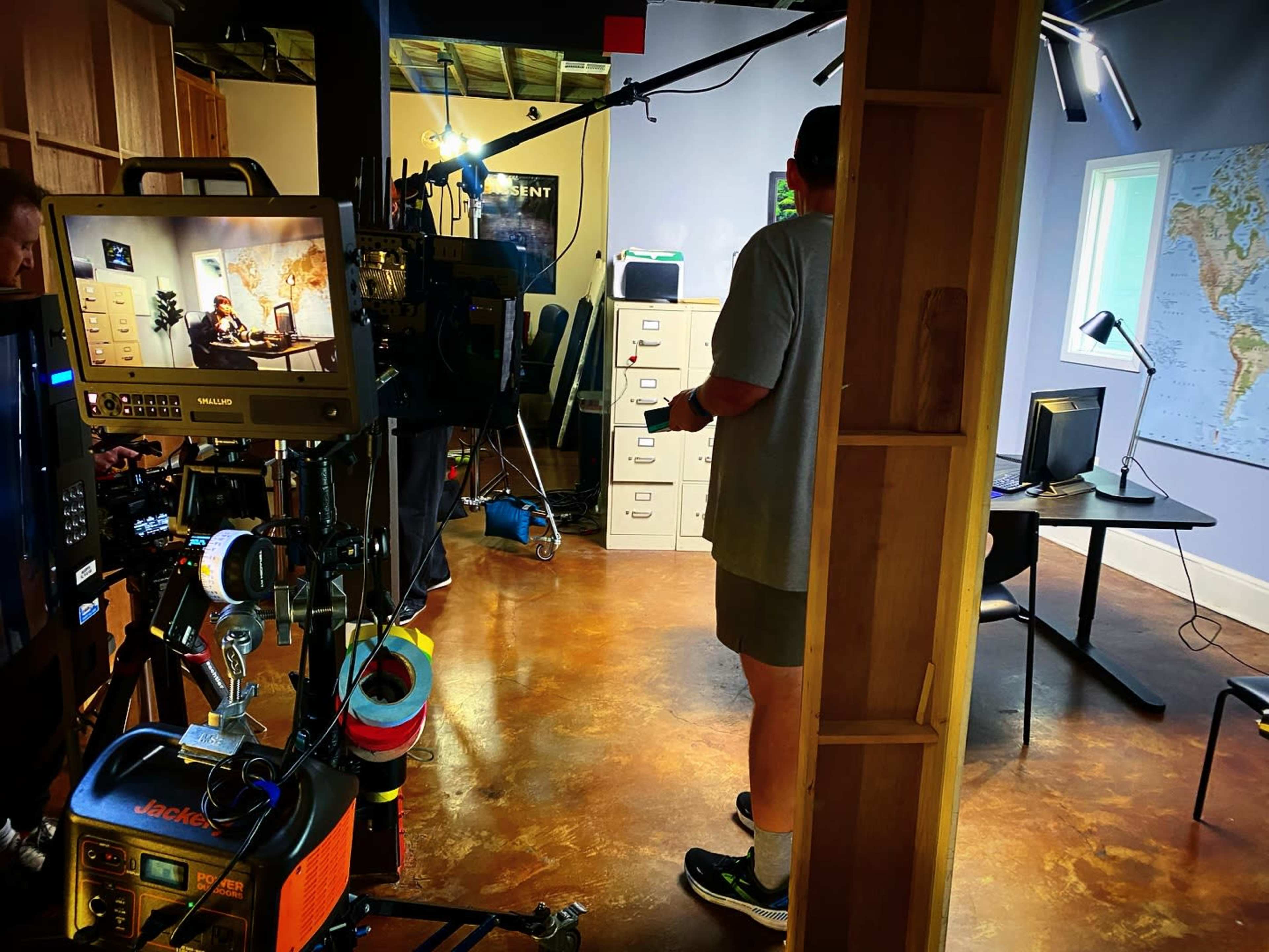 A film set features a crew member standing in a room equipped with cameras, lights, and various pieces of furniture.