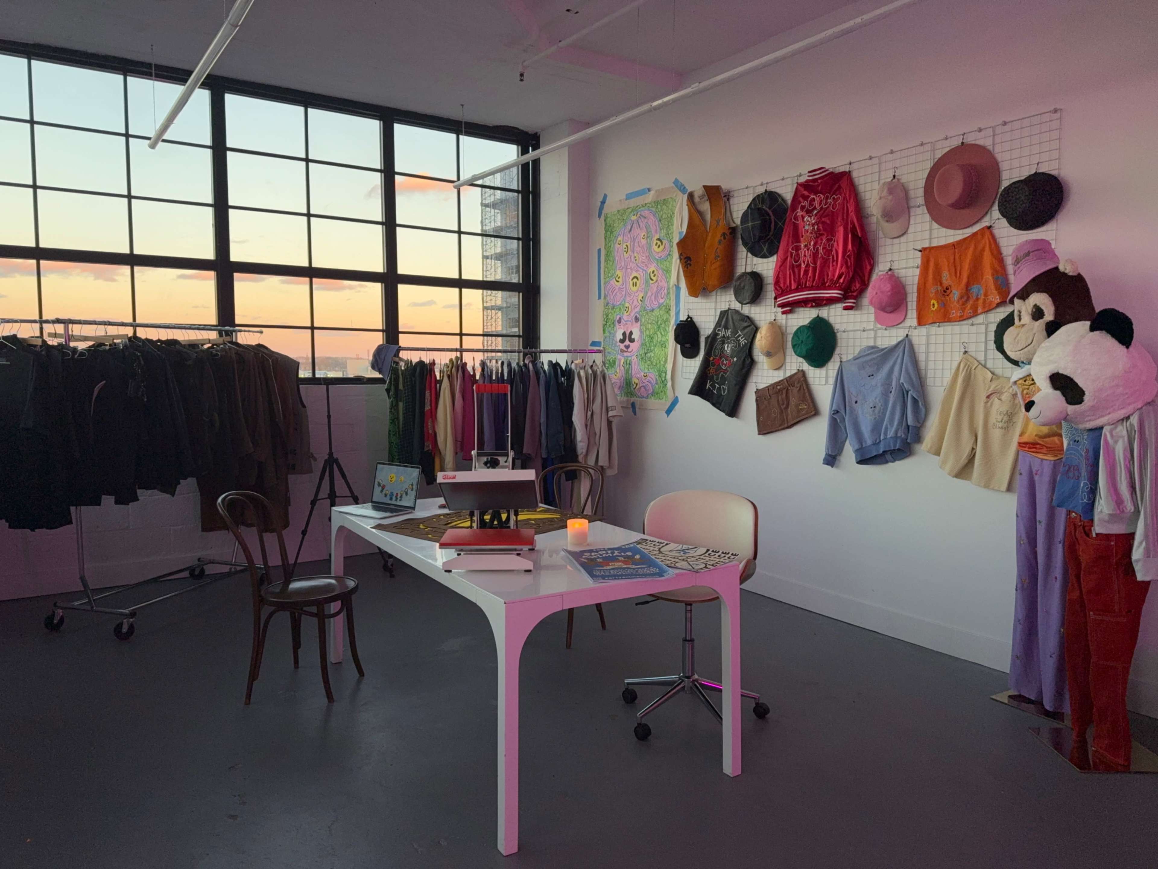 The image shows a bright, open studio space with a white table and chair, surrounded by racks of clothing and a wall displaying various garments and accessories, all against a backdrop of a large window revealing a sunset.