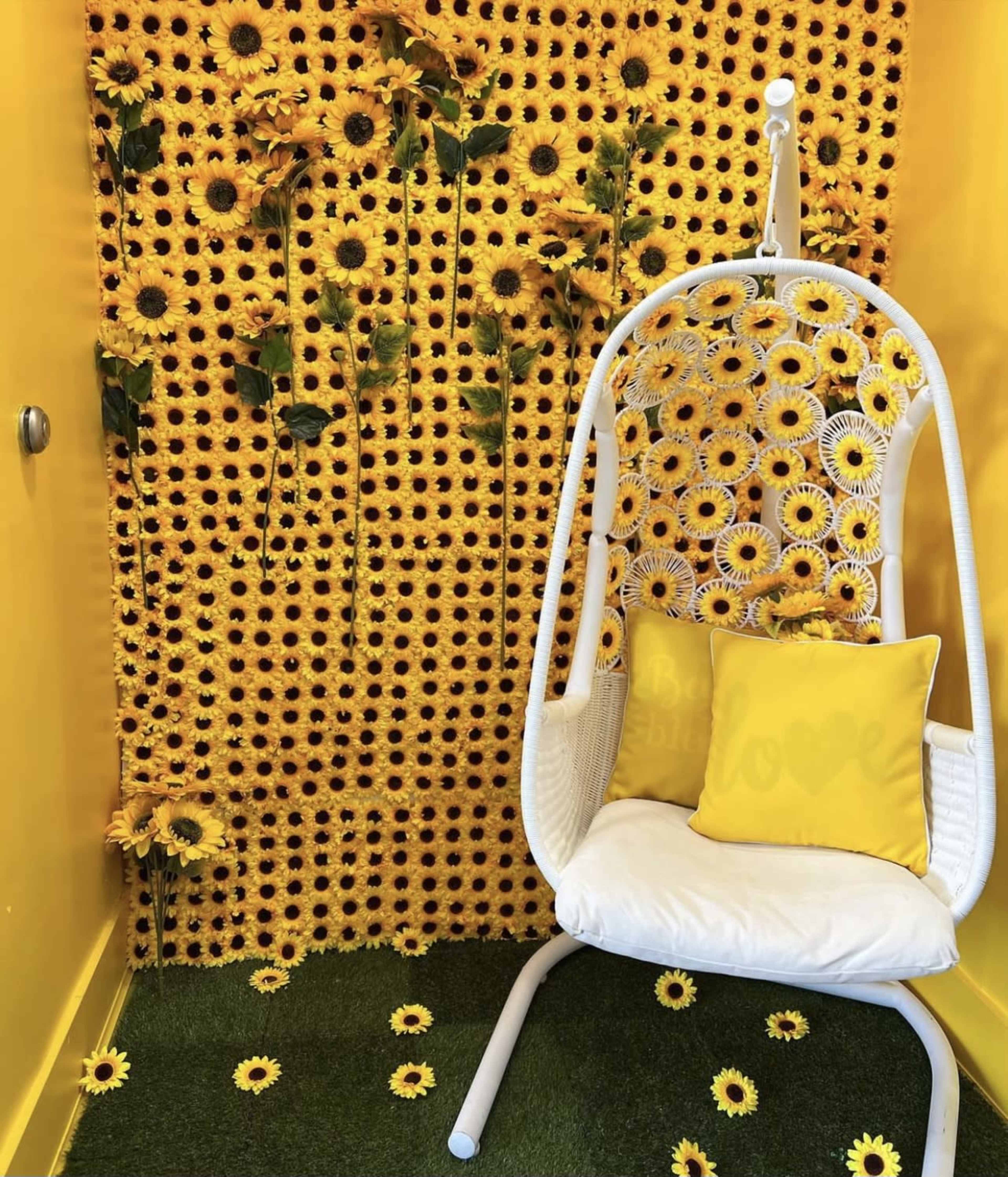 A yellow-themed room featuring a white hanging chair in front of a wall covered in sunflowers, with artificial grass and scattered sunflower decorations on the floor.
