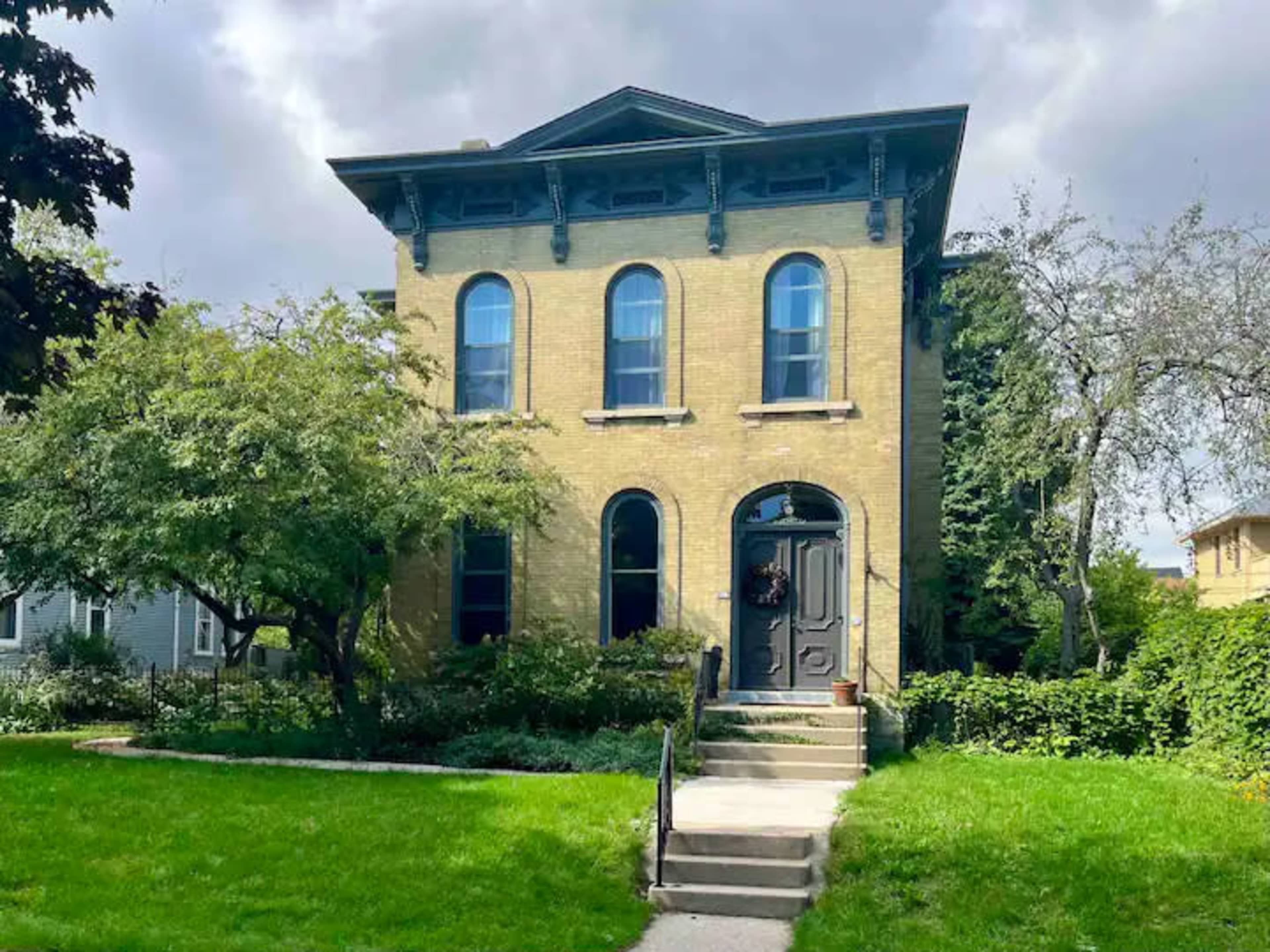 A historic two-story brick house features arched windows and a central front door, surrounded by greenery and a well-maintained lawn.