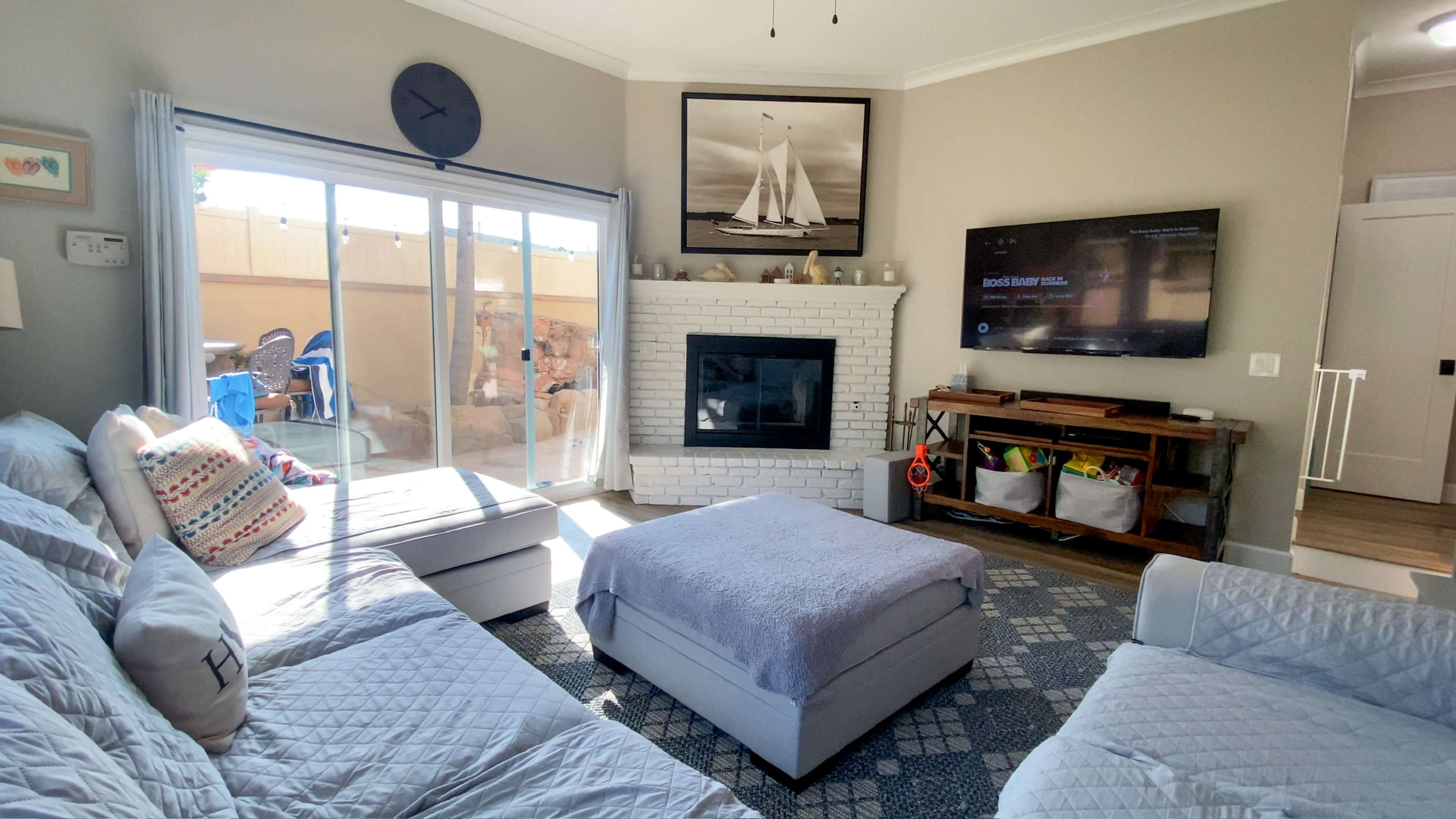 The living room features a white brick fireplace under a wall-mounted TV, with a large window overlooking an outdoor space, and seating arranged around a light-colored ottoman.