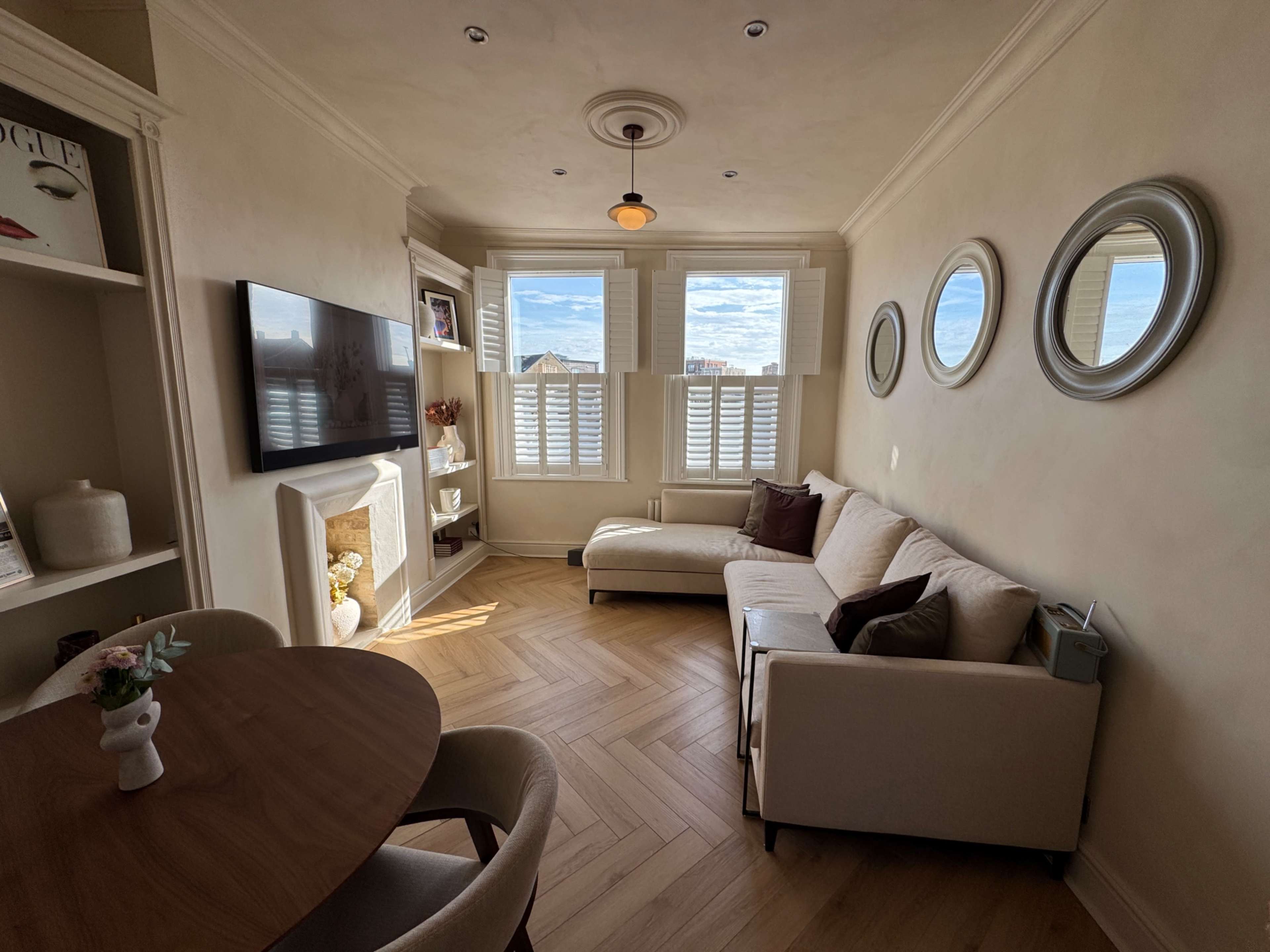 The image shows a bright living room with a sectional sofa, a round dining table, a television mounted on the wall, and windows covered with plantation shutters.