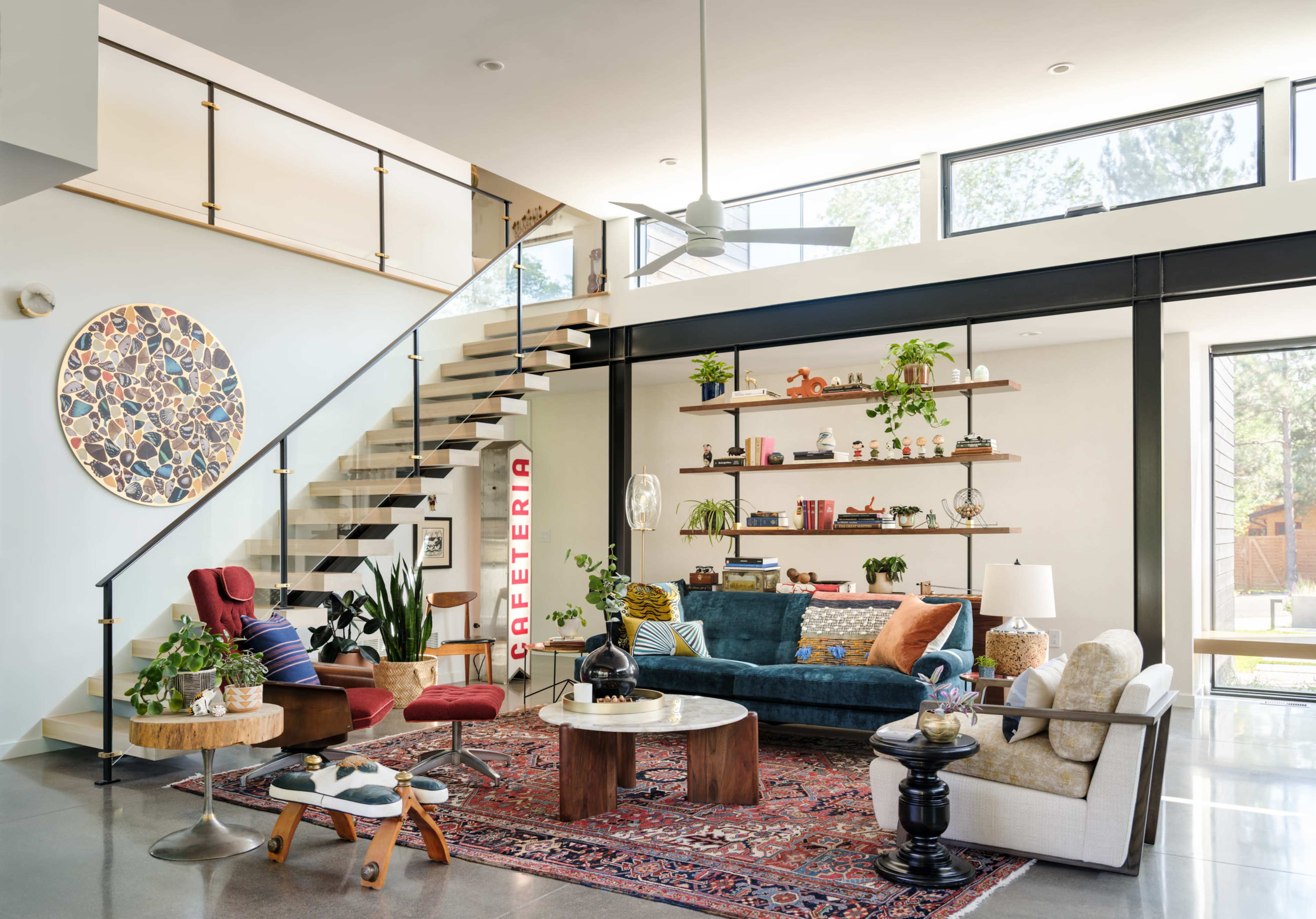 The image shows a modern living room with a staircase, various plants, a colorful rug, and a mix of seating options arranged around a coffee table.